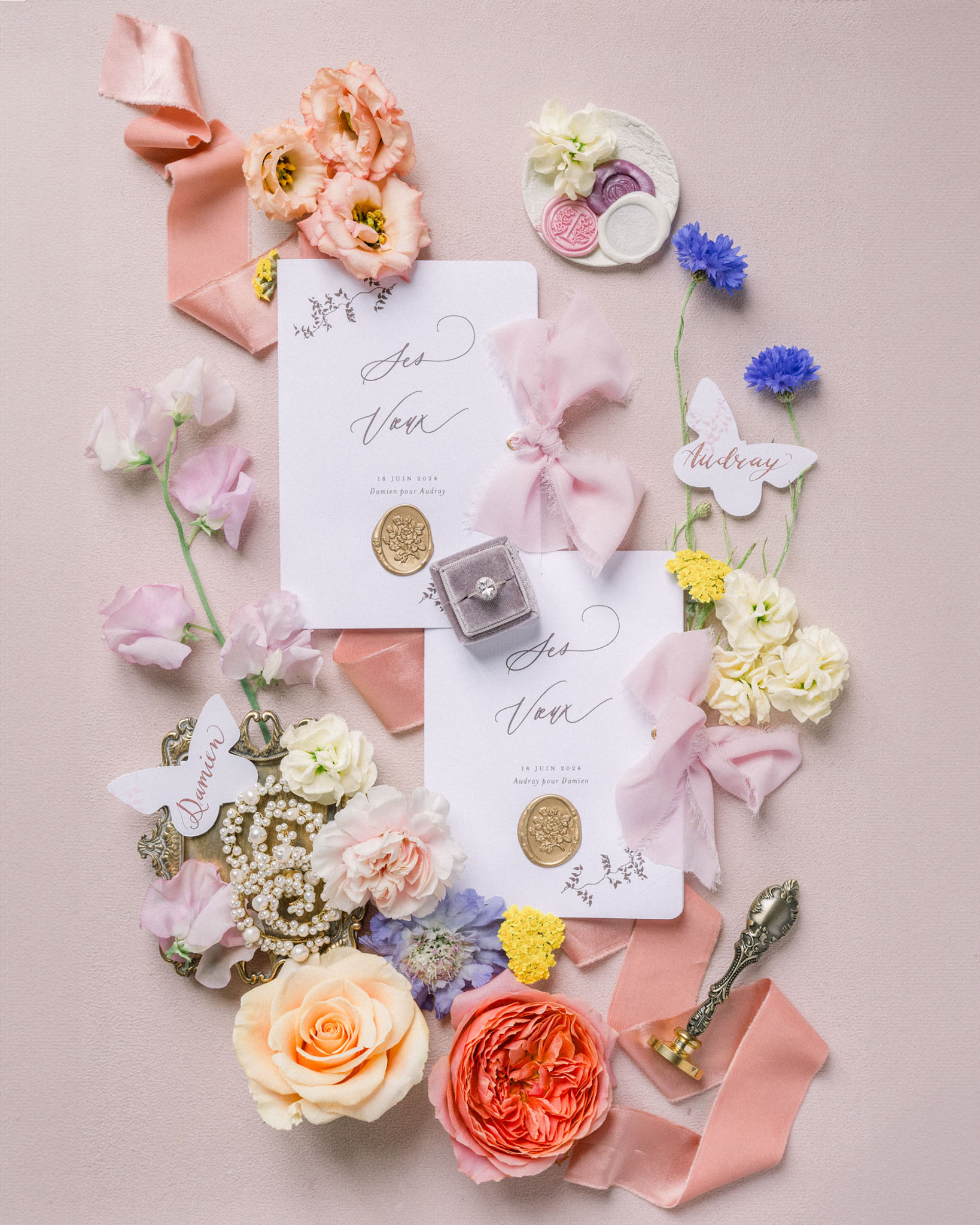 Flat lay of wedding invitations with gold wax seals, diamond ring, pink ribbons, peach roses and coral peonies