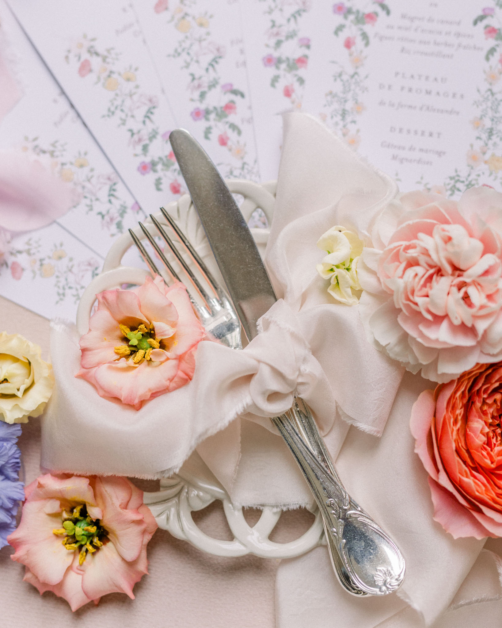 Place setting with ornate silver flatware, cream napkin with silk bow, and coral and blush peonies