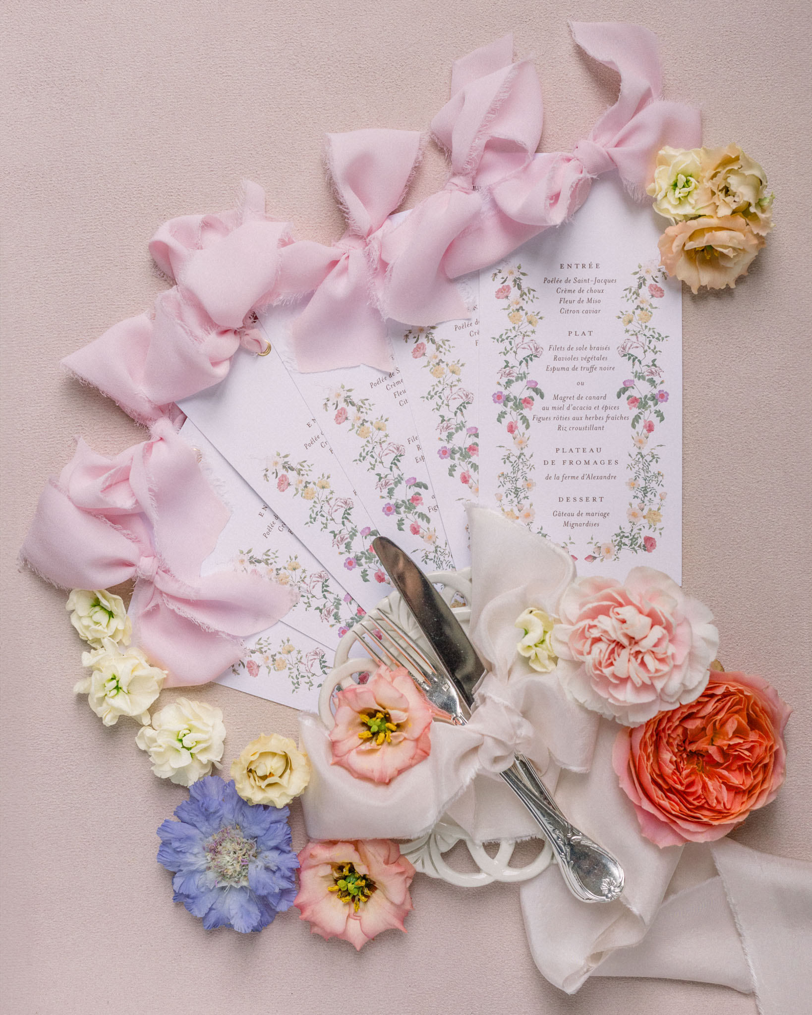 Flat lay of French wedding menu card with pink peonies, cream roses, vintage silverware, and silk ribbons