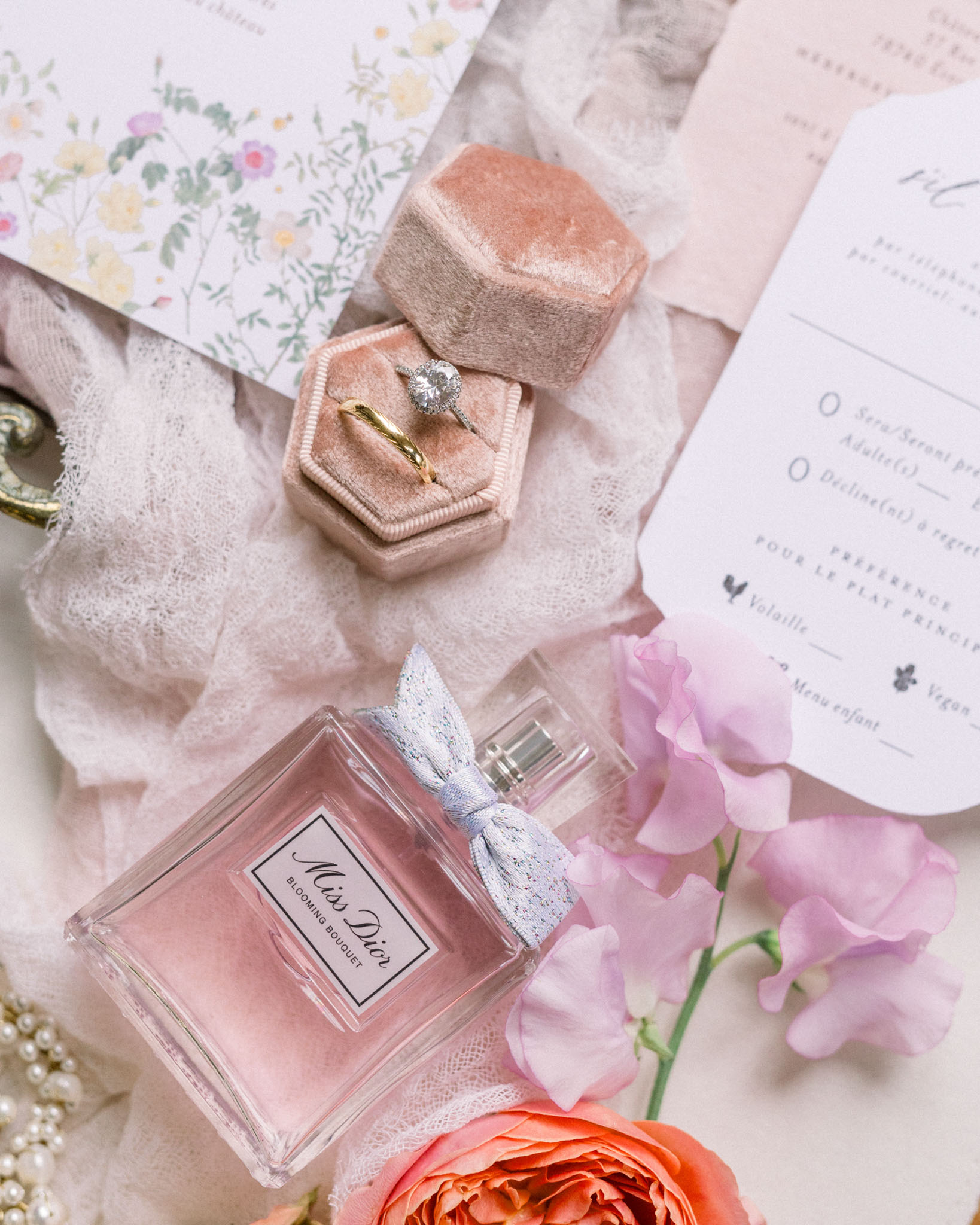 Flat lay of bridal accessories with engagement ring in dusty rose velvet box, perfume, and floral stationery on blush linens