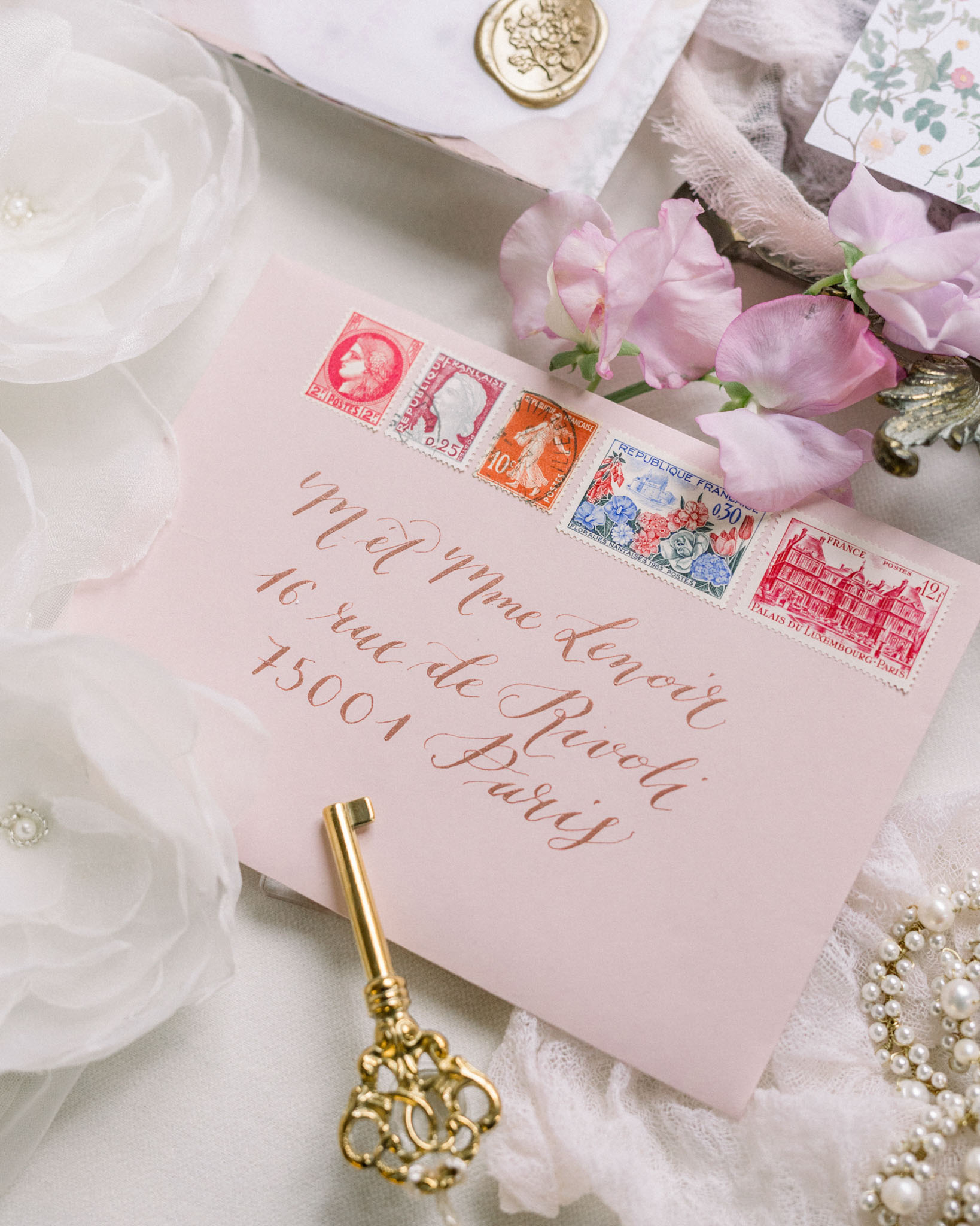 Flat lay of wedding stationery with pink envelope, copper calligraphy, French stamps, gold wax seal, and peonies