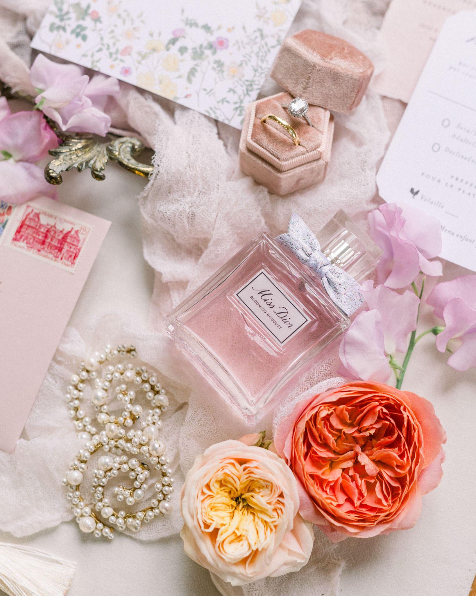 Flat lay of velvet ring box with diamond ring, Miss Dior perfume, pink roses, coral peonies, and floral stationery