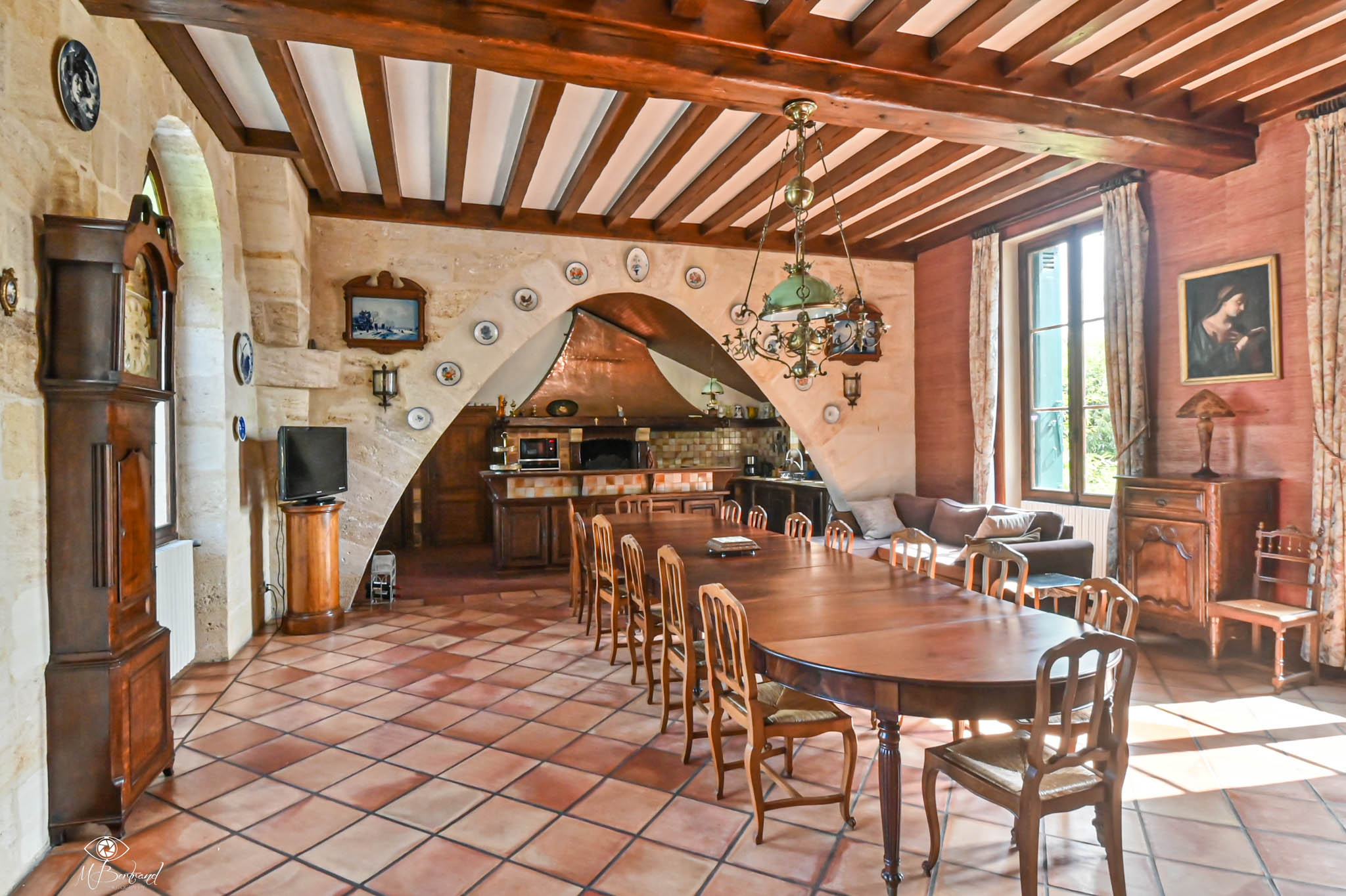 Rustic French dining room with beam ceiling oval table rush chairs grandfather clock and stone arch kitchen