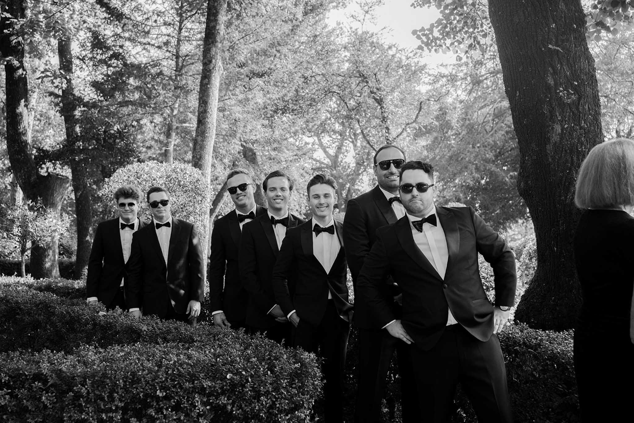 Black and white group portrait of groom and groomsmen in tuxedos in wooded garden setting