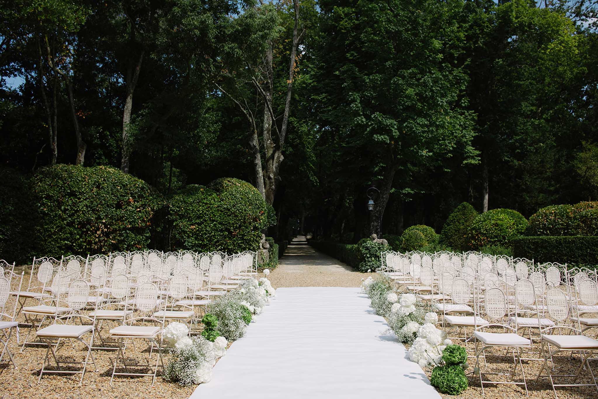 Outdoor ceremony setup with white Chiavari chairs and hydrangea aisle markers in a formal topiary garden