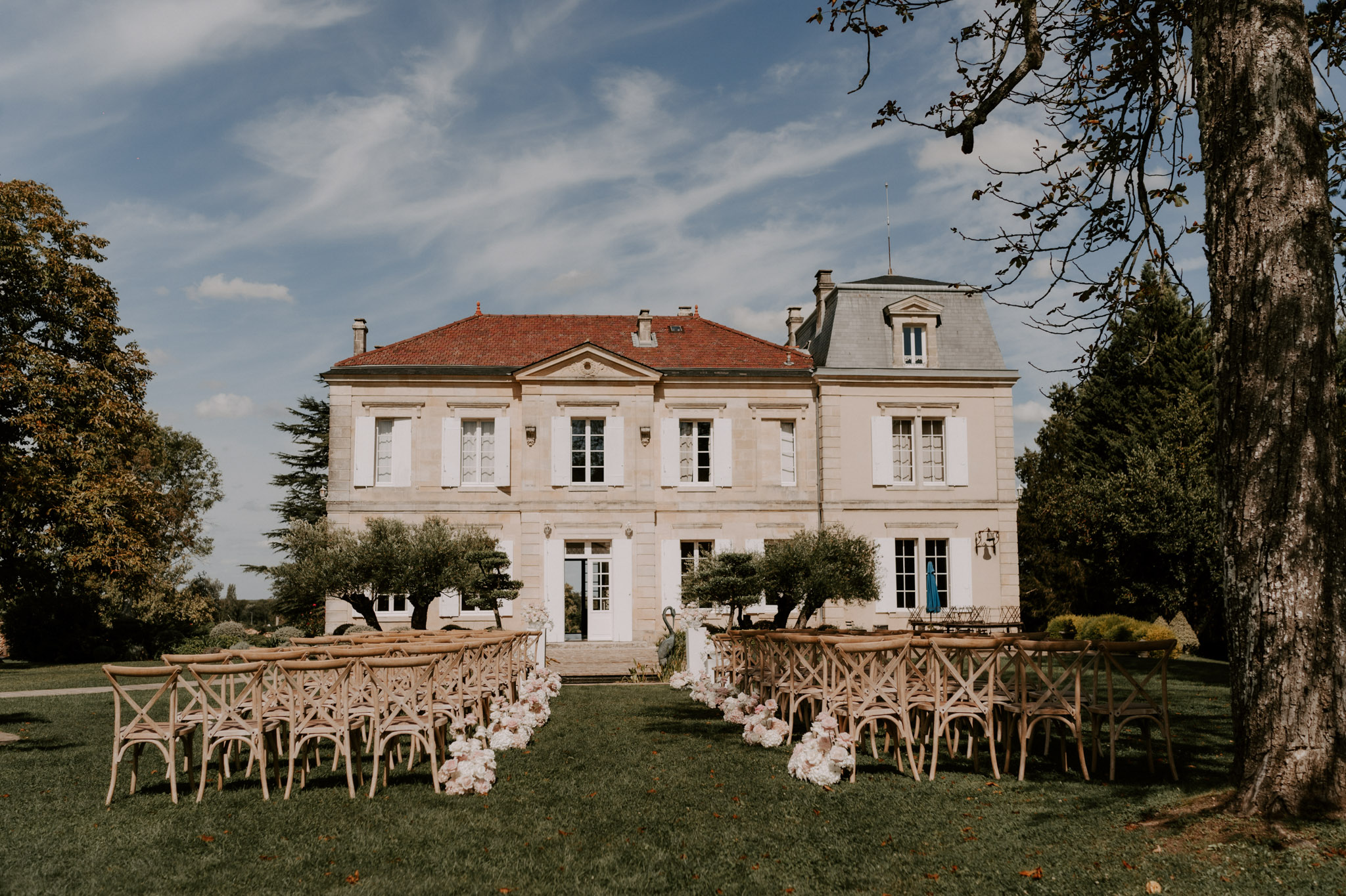 Outdoor ceremony setup with wooden cross-back chairs and blush floral aisle markers facing a limestone chateau