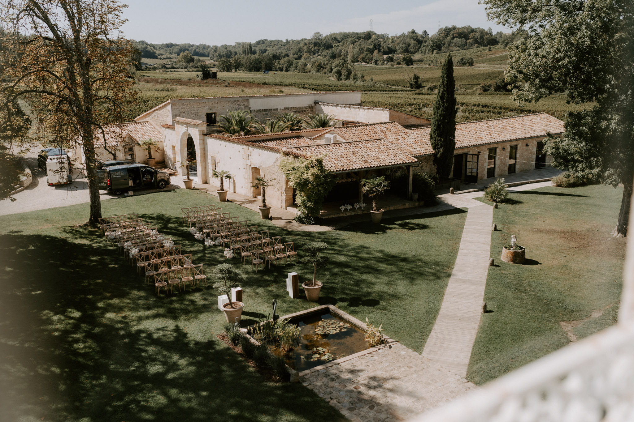 An aerial wide shot taken from an elevated position overlooking a French wine estate venue set up for an outdoor wedding ceremony. The grounds feature rows of natural wood cross-back chairs arranged in a curved semicircular configuration on a lawn, with white floral accents at the ends of select rows. A rectangular ornamental pond with lily pads sits in the foreground, flanked by a stone-paved walkway. The venue building is a low-lying stone structure with terracotta roof tiles, arched doorways, and a covered terrace, surrounded by potted palm trees and cypress trees. Vineyard rows are visible in the background extending across the hillside. The overall decor style is rustic-natural with warm tones. Potential venue feature image.