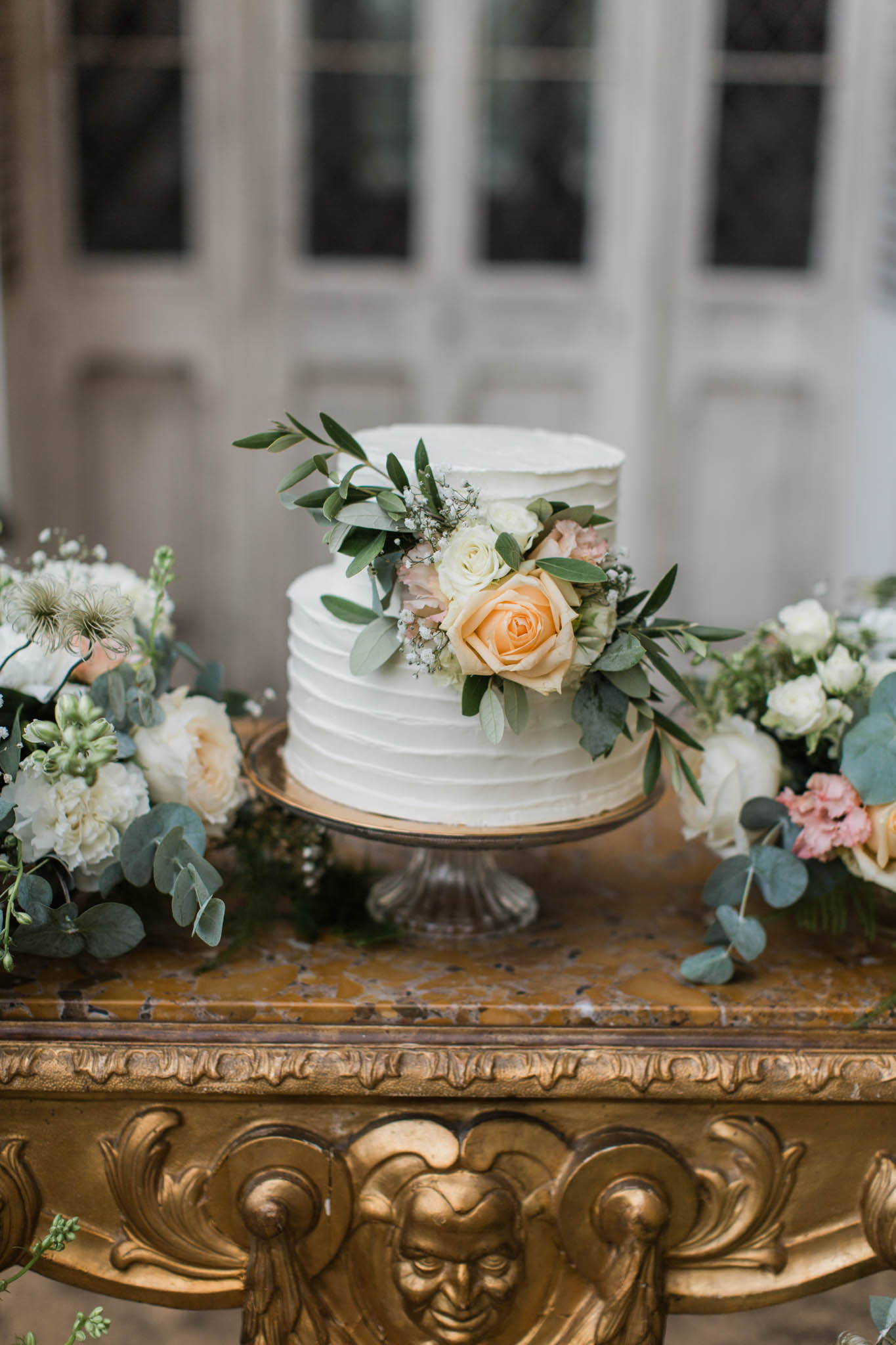 Two-tier white buttercream wedding cake with fresh peach roses and eucalyptus on gilded console table