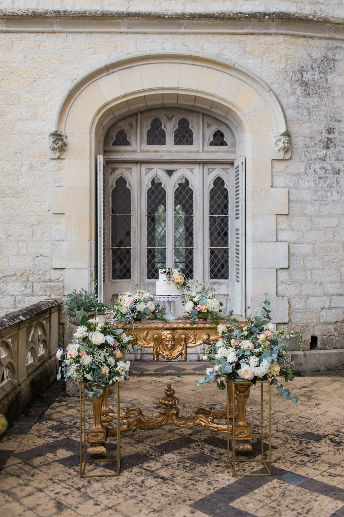Two-tiered white and gold wedding cake flanked by cascading ivory peony arrangements in a Gothic stone courtyard