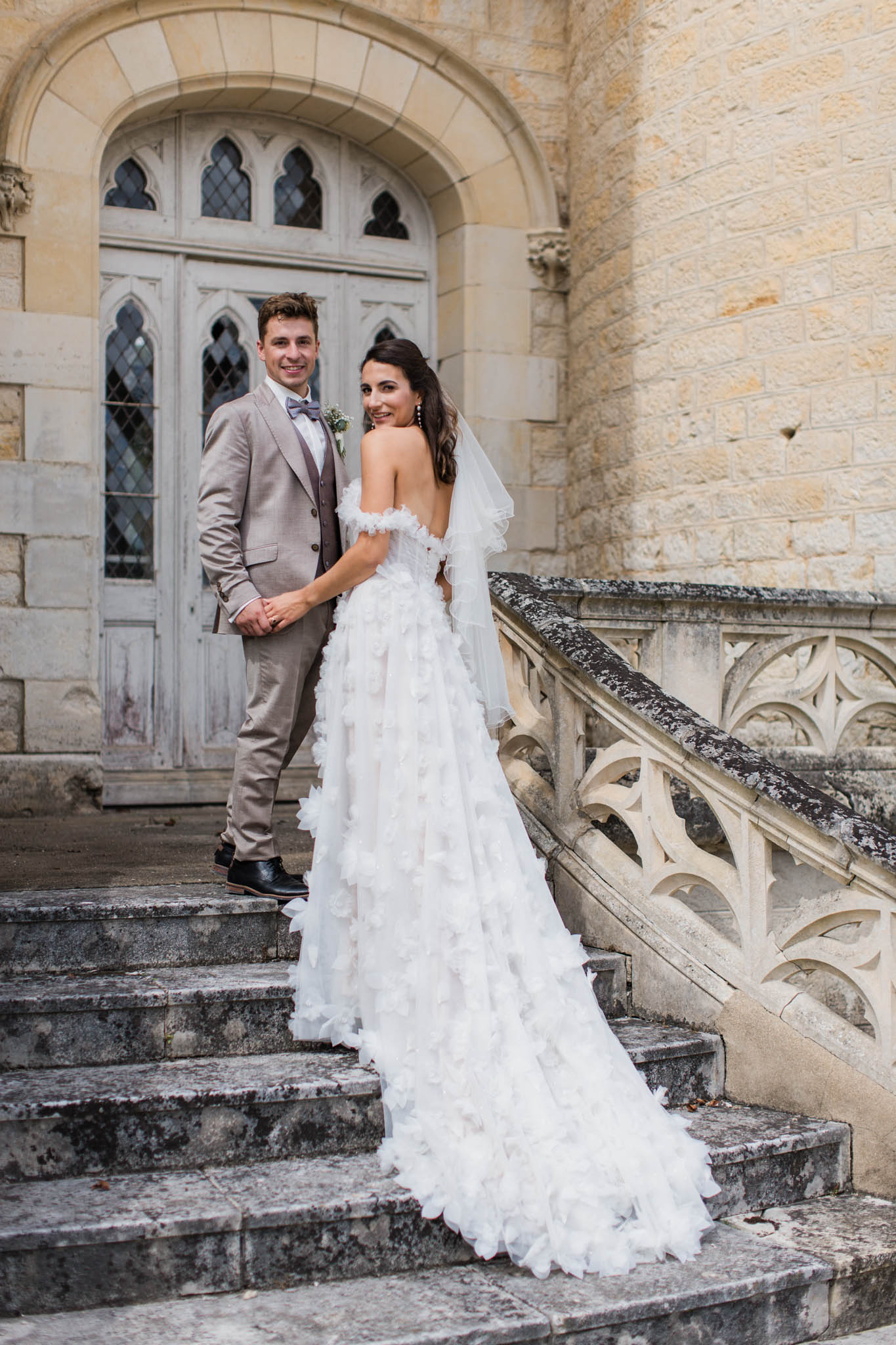 Bride in ruffled-tier off-shoulder gown and groom in taupe linen suit posing on Gothic chapel steps after ceremony