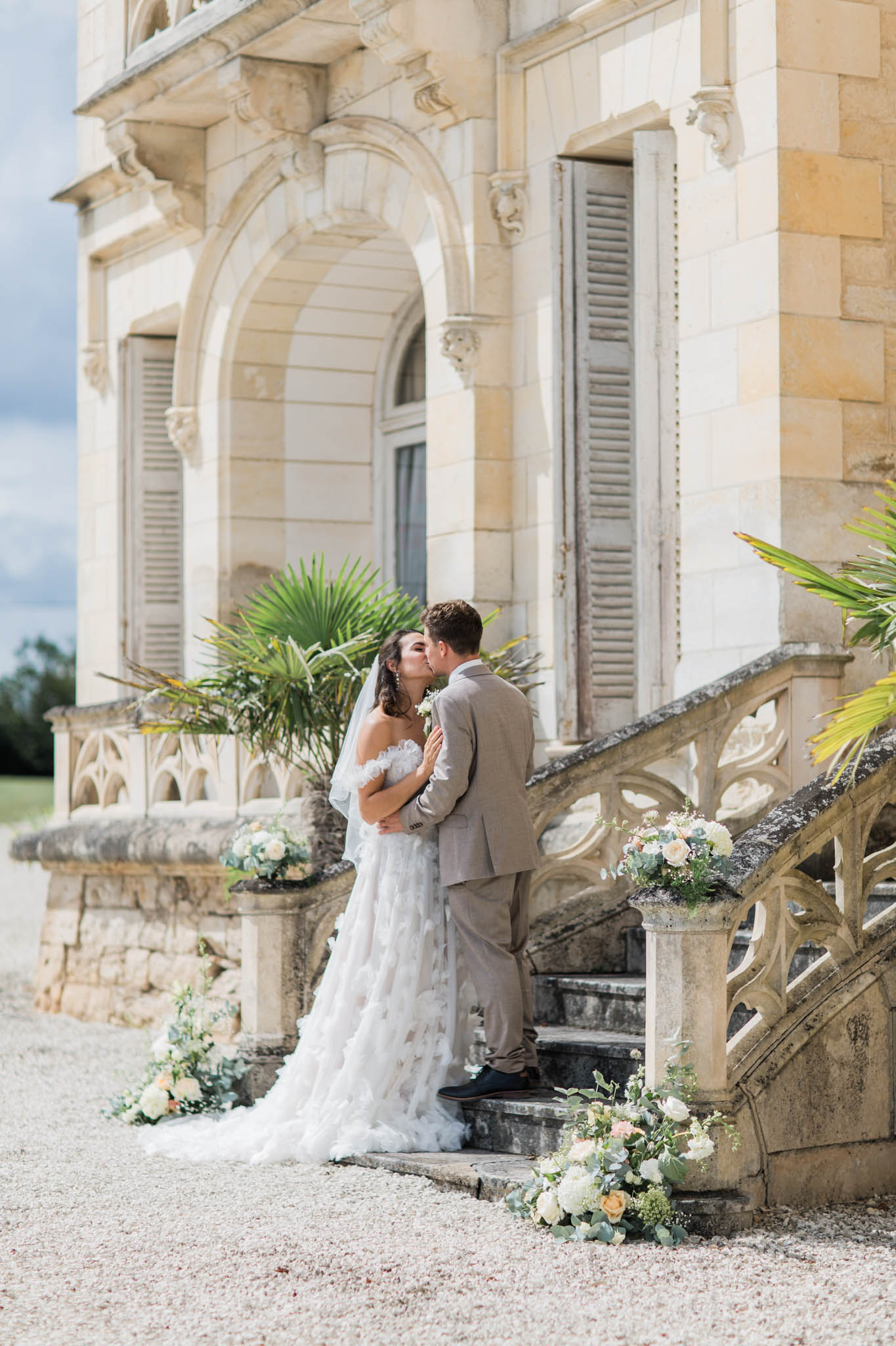 Bride and groom embracing on chateau stone steps with cream and blush floral arrangements and fan palms