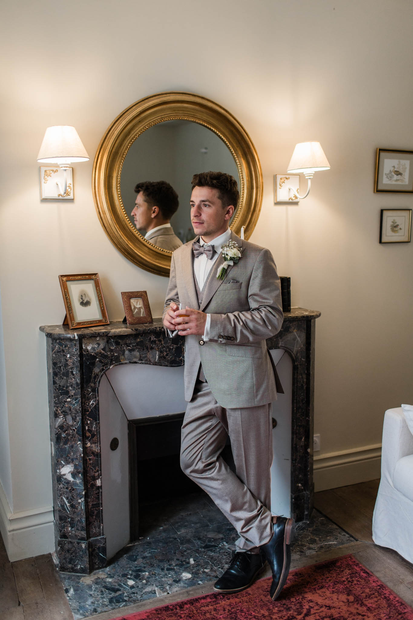 Groom in gray-beige suit with lavender bow tie posing beside a dark marble fireplace with gold-framed circular mirror above.