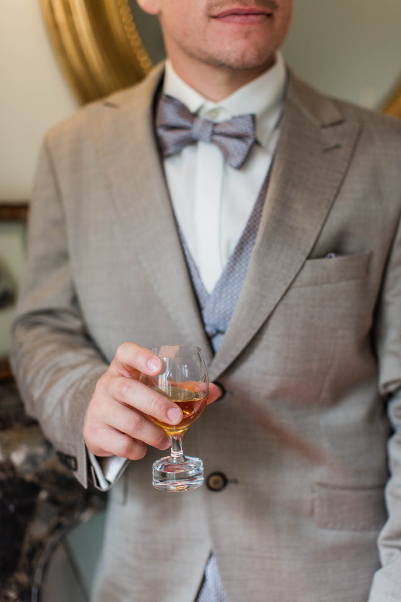 Close-up of groom holding amber drink wearing grey linen suit and slate blue bow tie
