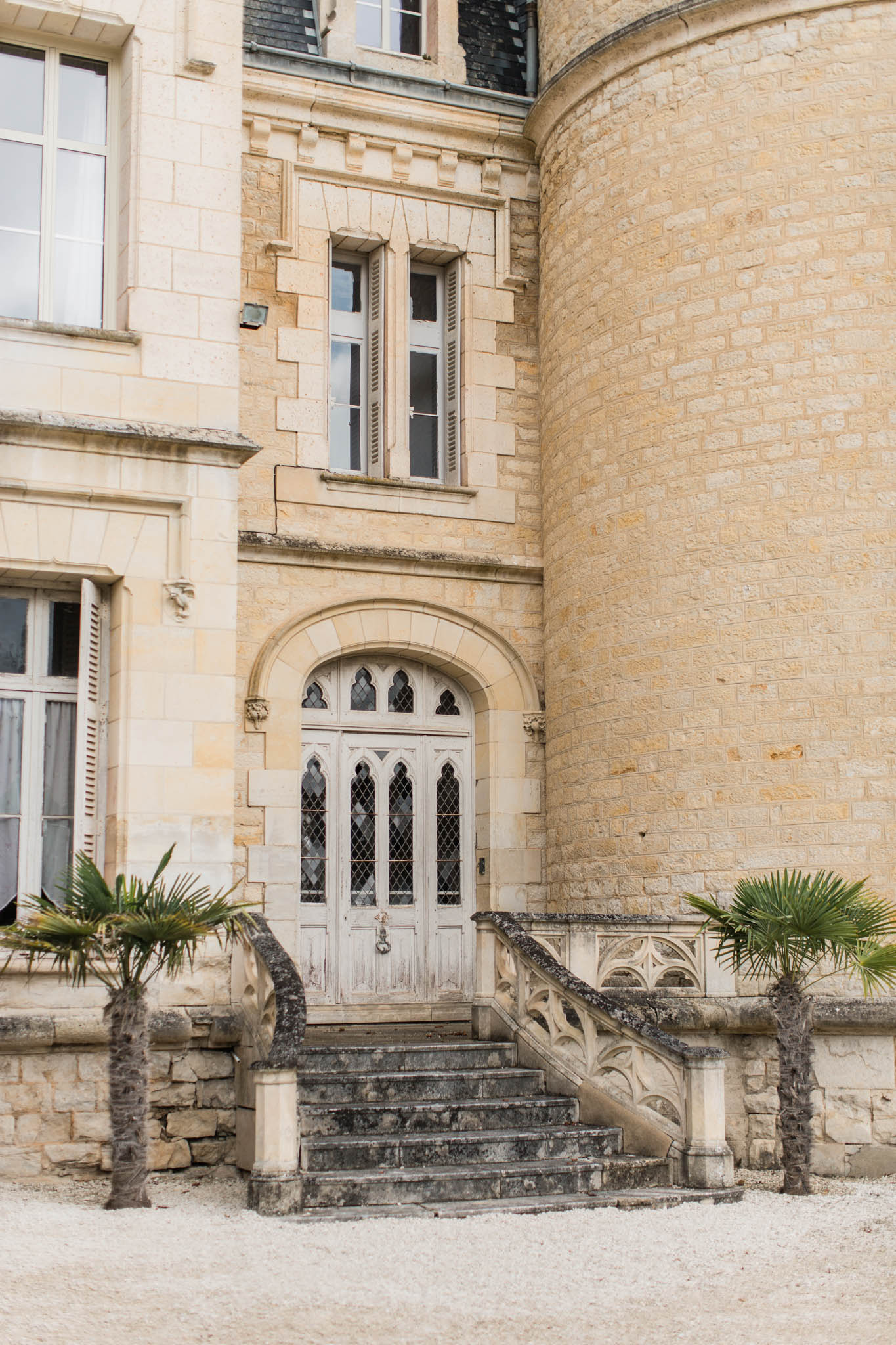 Historic stone chateau entrance with Gothic arched doorway, potted palms, and cylindrical tower