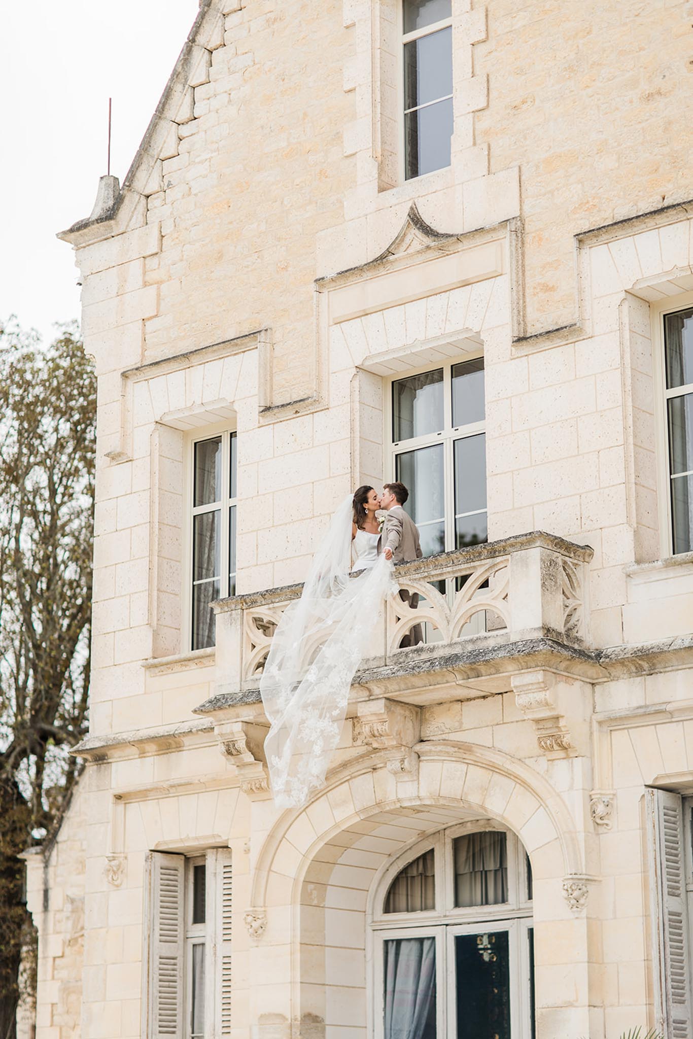 Groom kisses bride's cheek on stone balcony of classical cream château, her white veil flowing behind them