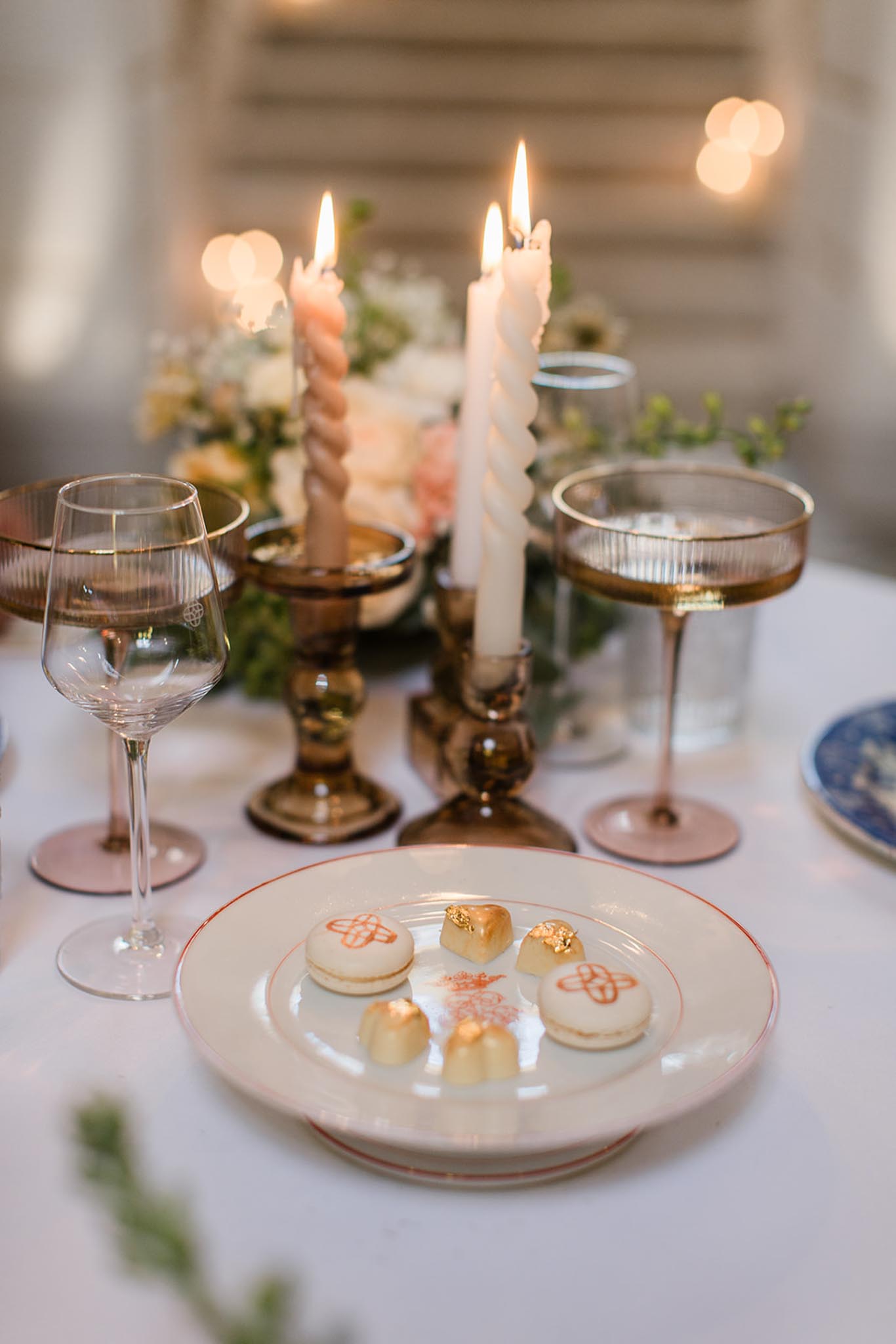 White plate with rose-gold rim holding macarons and petit fours at wedding reception, brass candelabras behind