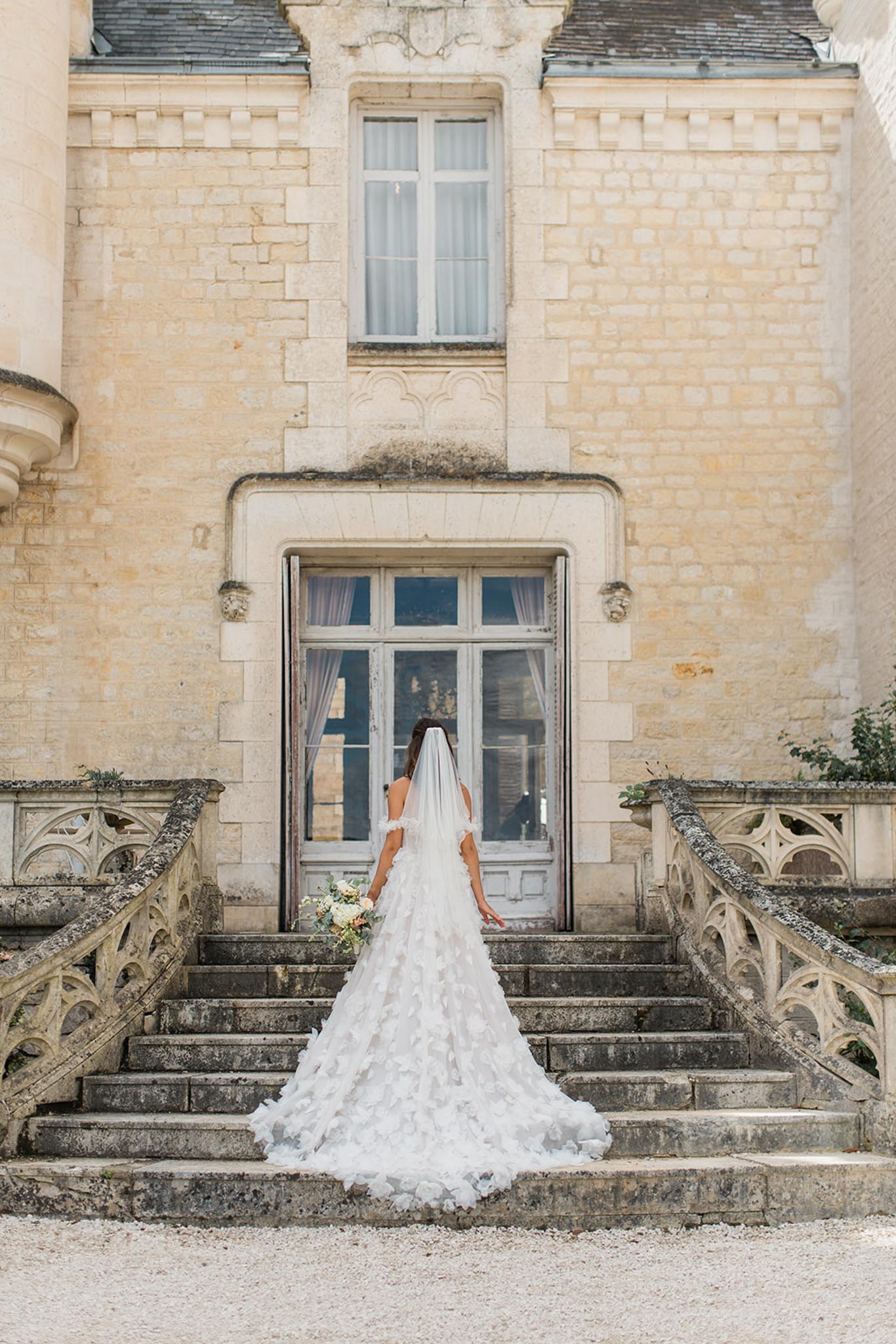 Bride from behind in a white layered gown with cathedral veil trailing down the stone steps of a classical French château entrance.