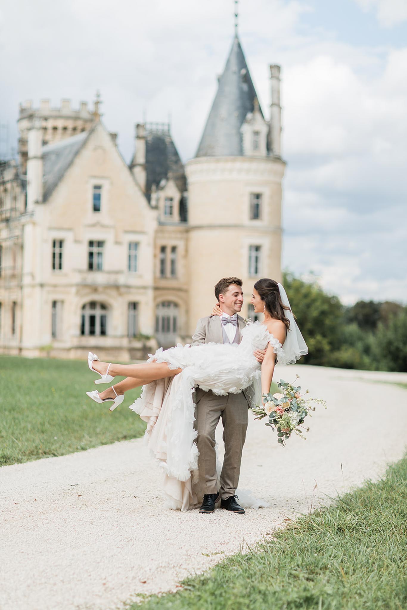 Groom carrying bride on château grounds, bride in white ruffled gown with château turret visible behind them