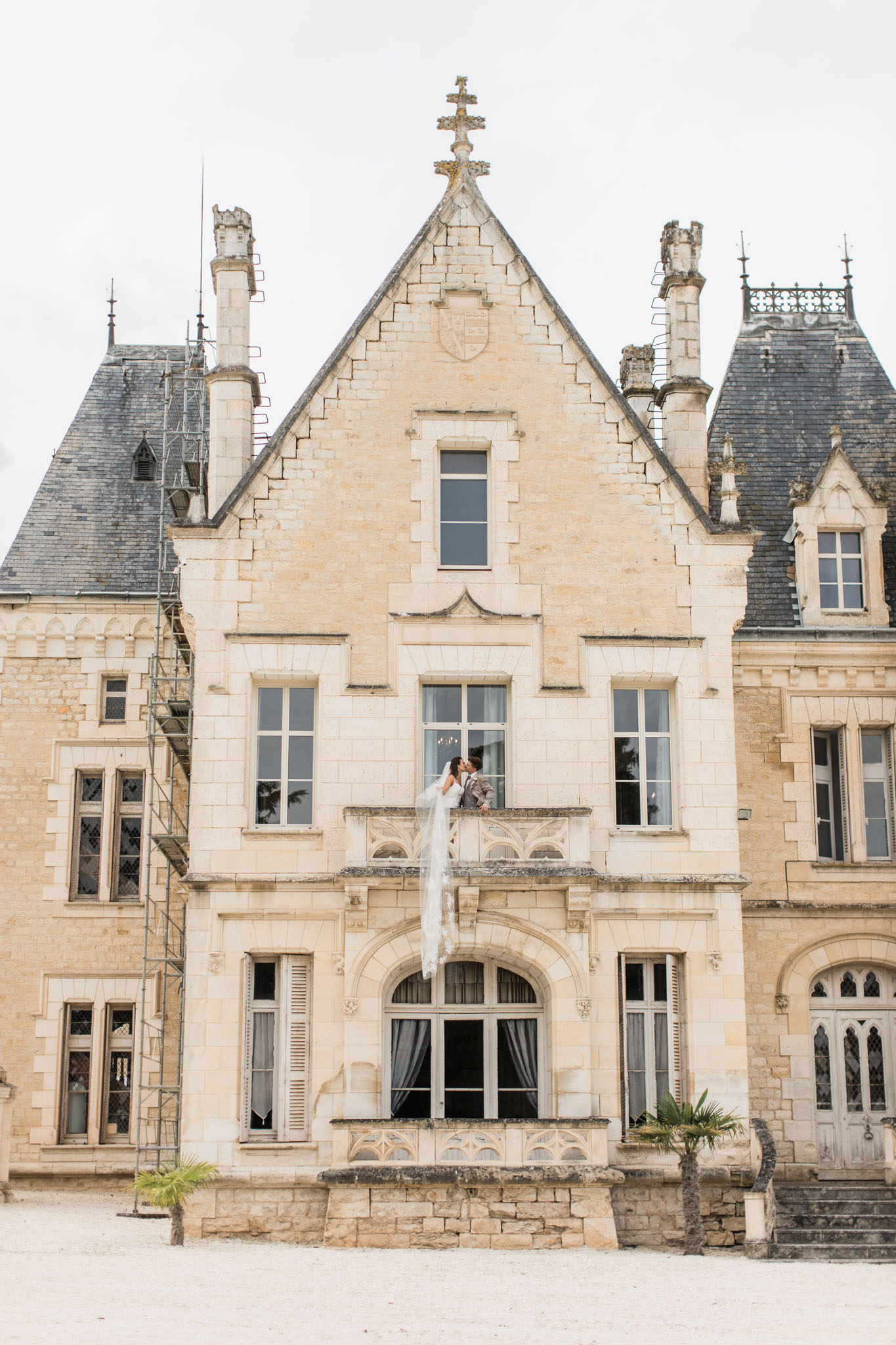 Bride and groom standing on stone balcony of grand château with arched windows and crenellated towers