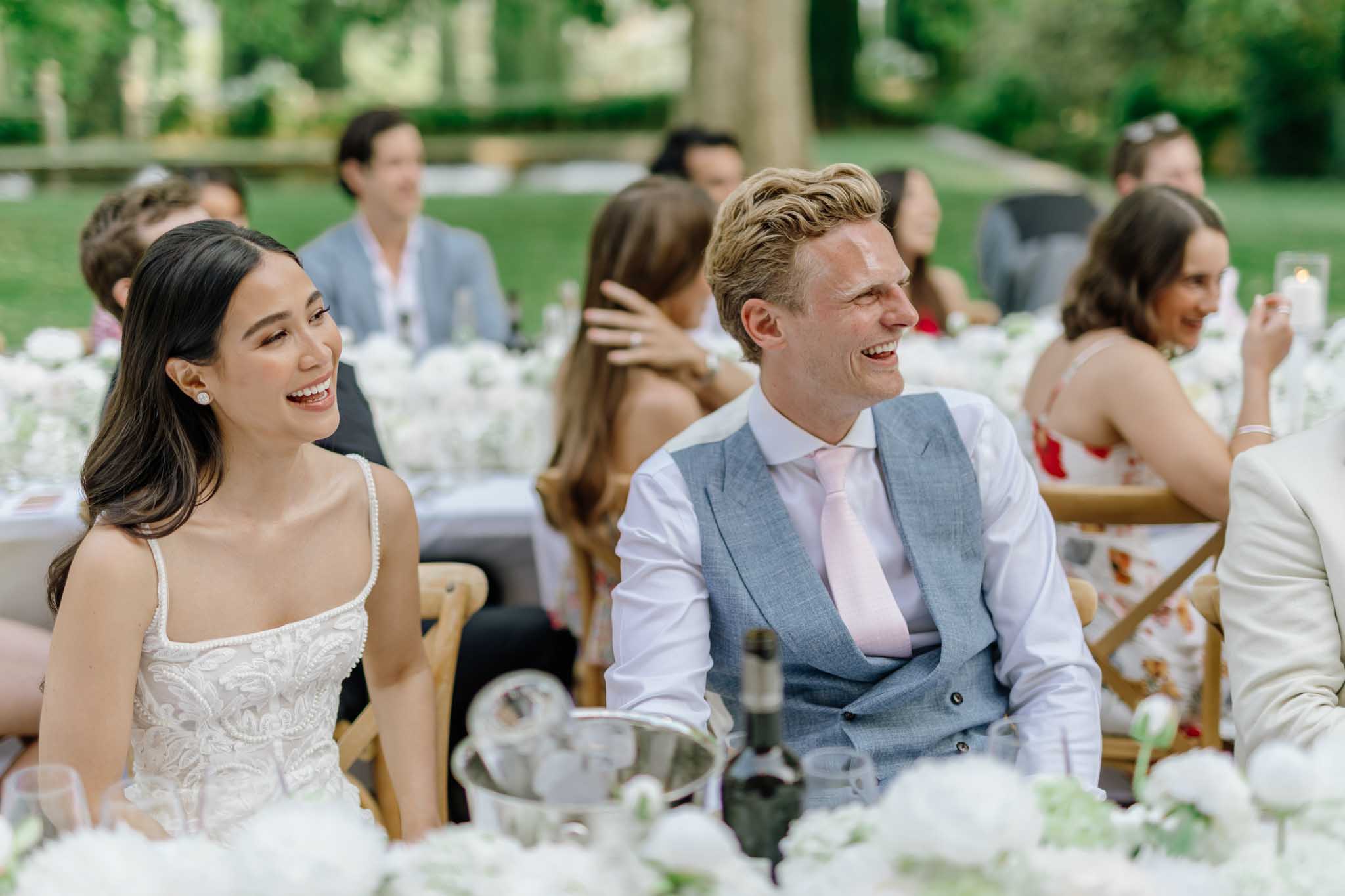 Bride and groom laughing at a long reception table with white hydrangea centerpieces in an outdoor garden setting