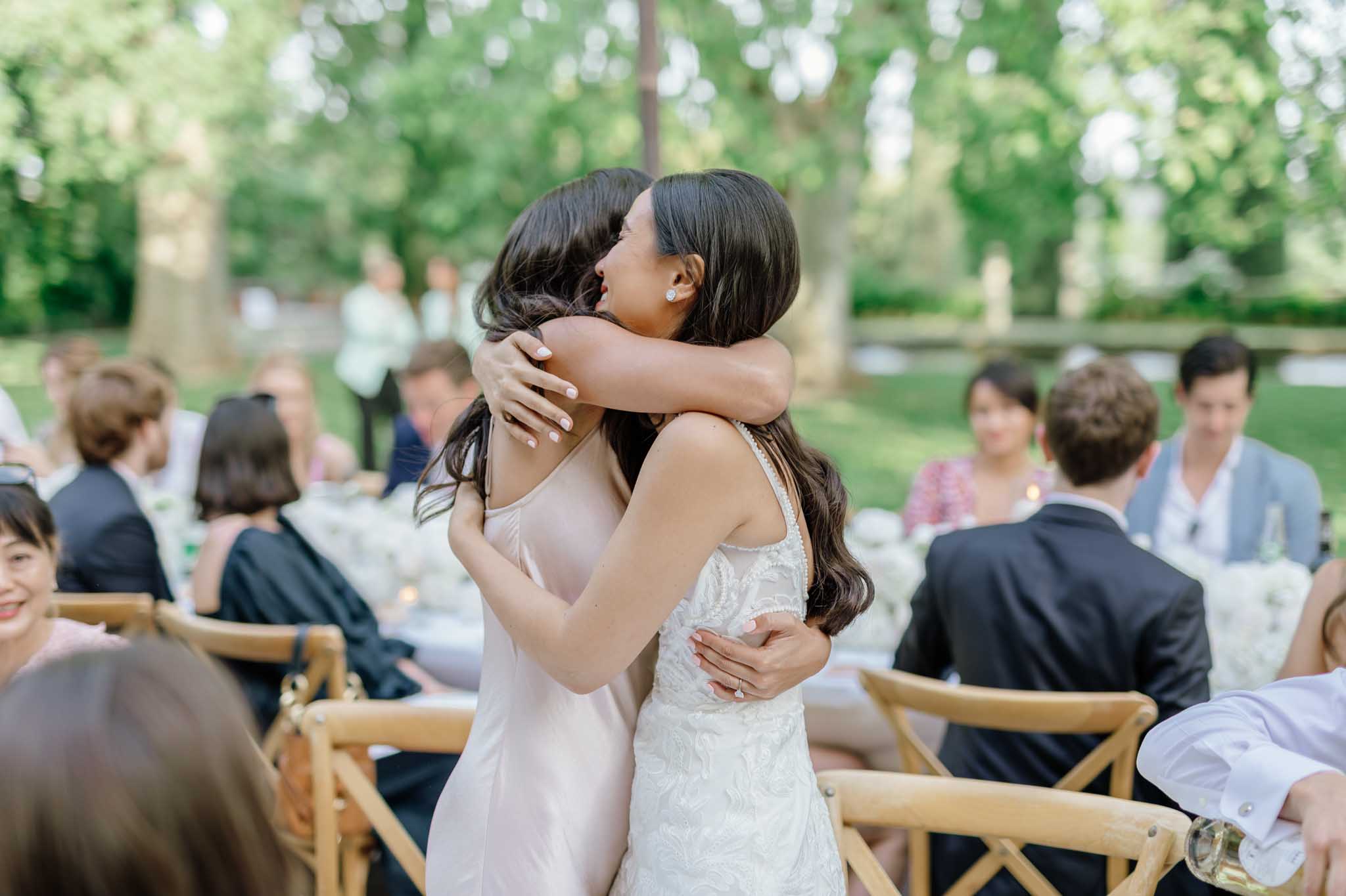 Bride in white lace gown embracing a guest in blush dress at outdoor garden reception