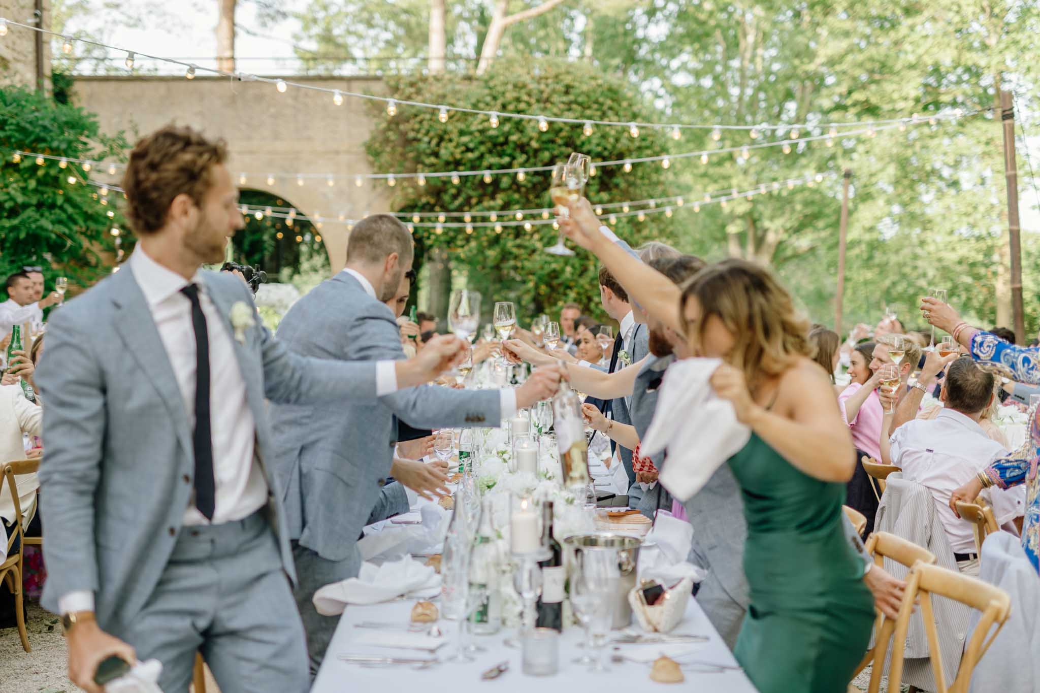 Guests raising glasses for toast at outdoor banquet table with string lights, ivy-covered walls, approximately 30-40 people