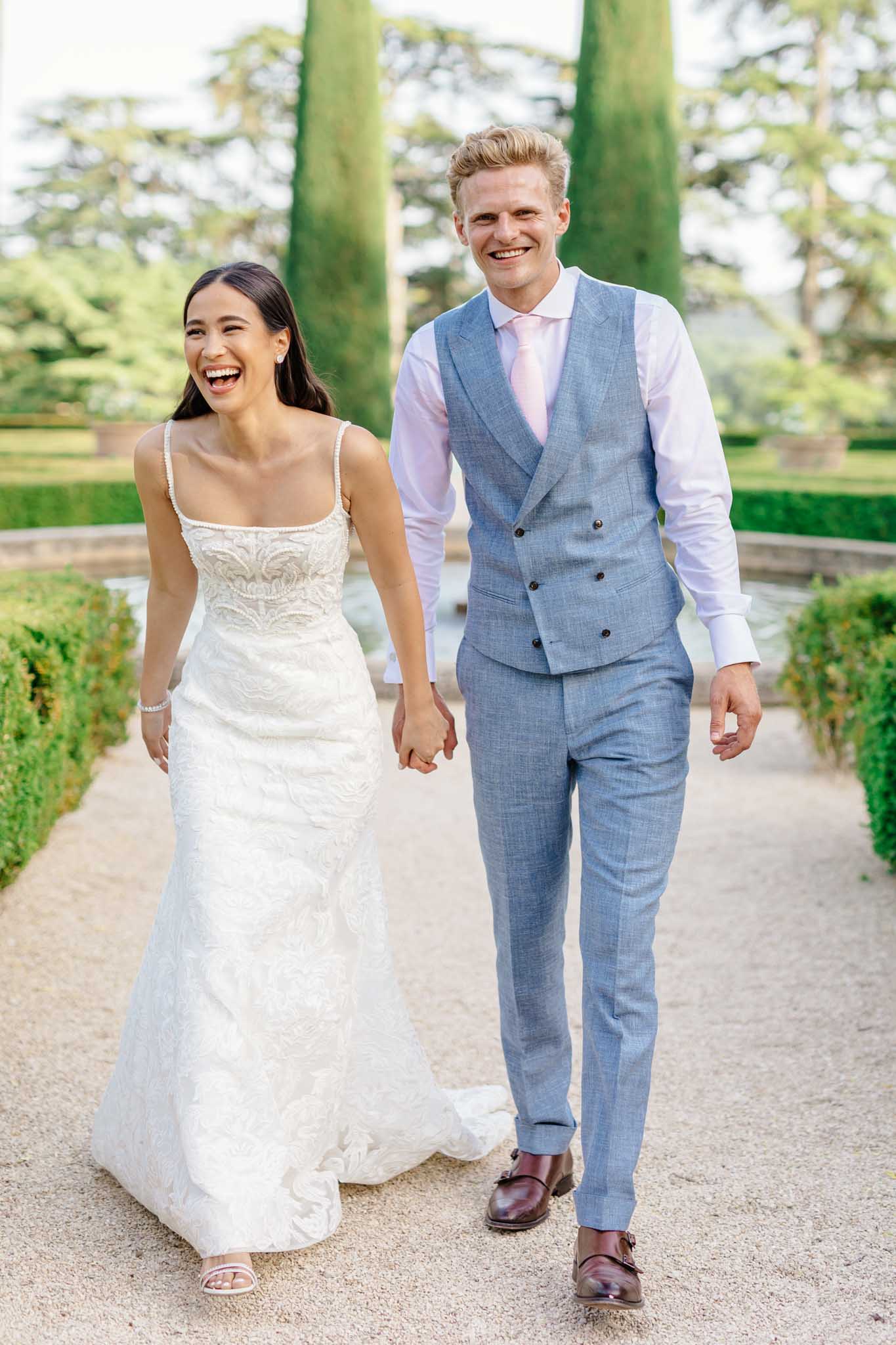Bride in ivory beaded mermaid gown walking with groom in light blue waistcoat along cypress-lined garden path