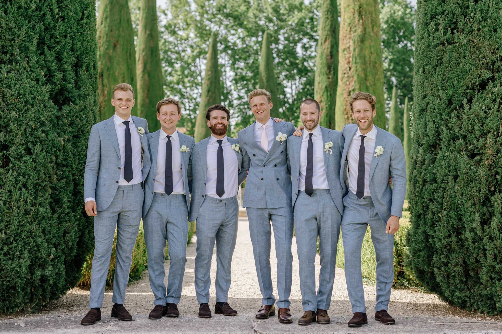 Groom and five groomsmen in coordinating blue-gray suits lined up between cypress trees