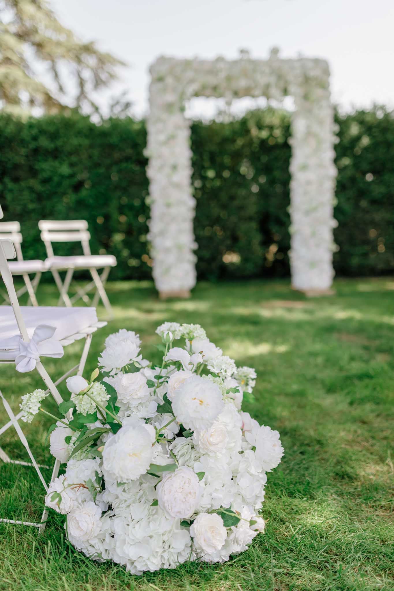 White peonies and roses bouquet on grass beside Chiavari chair with floral arch and hedges in garden ceremony