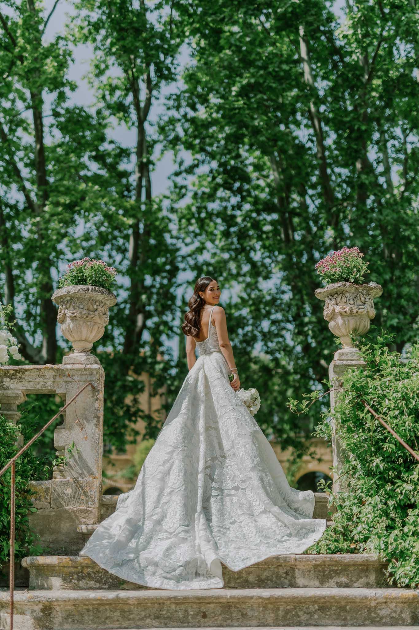 Bride in ivory lace ballgown standing between stone urns on a formal garden terrace with cypress trees