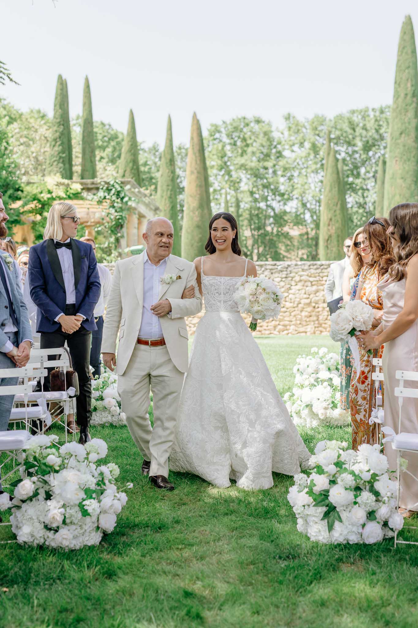 Bride walking down the aisle with her father through an outdoor garden ceremony lined with cypress trees