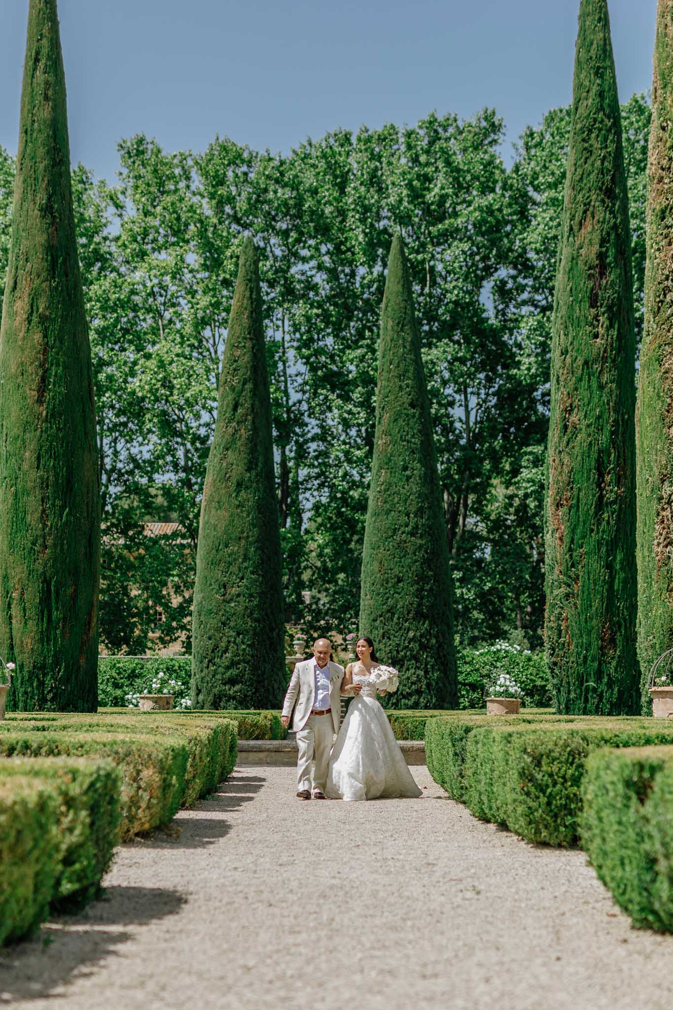 Bride in white ball gown and groom in beige suit walking down cypress-lined garden pathway