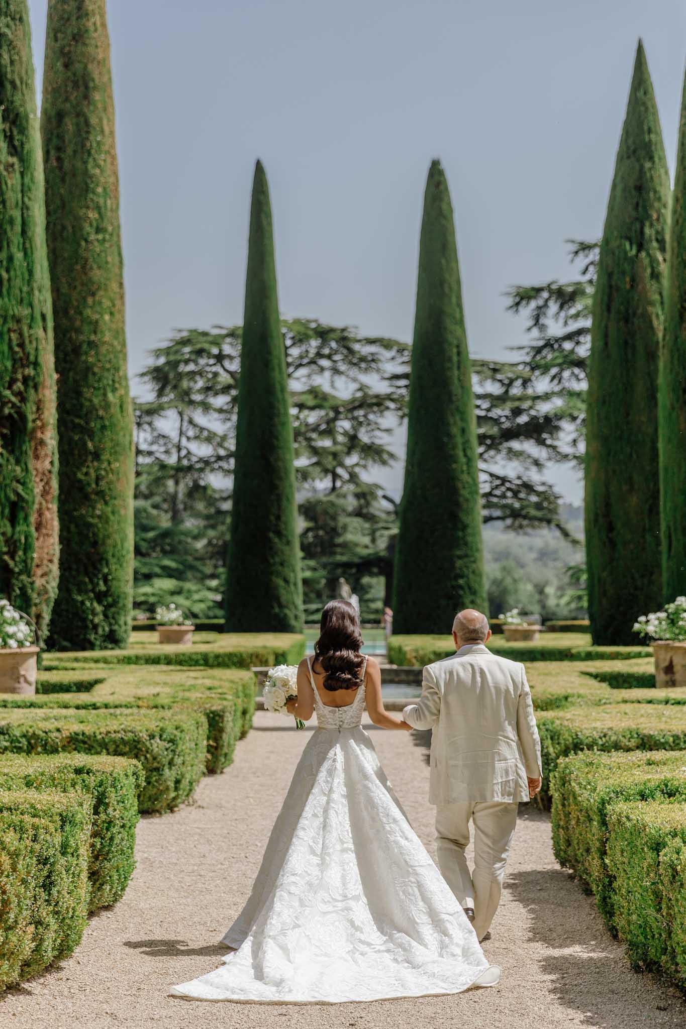 Bride in ivory ball gown and groom in cream suit walk formal Italian garden with cypress trees, viewed from behind