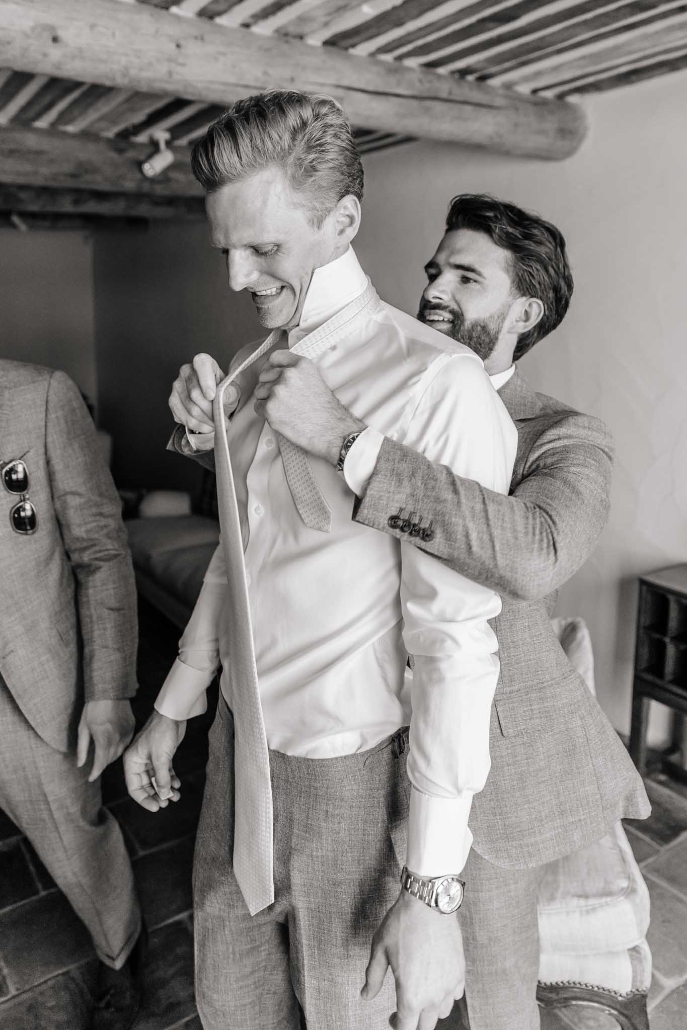 Black and white photo of groomsman helping groom with tie in venue with exposed wooden beams