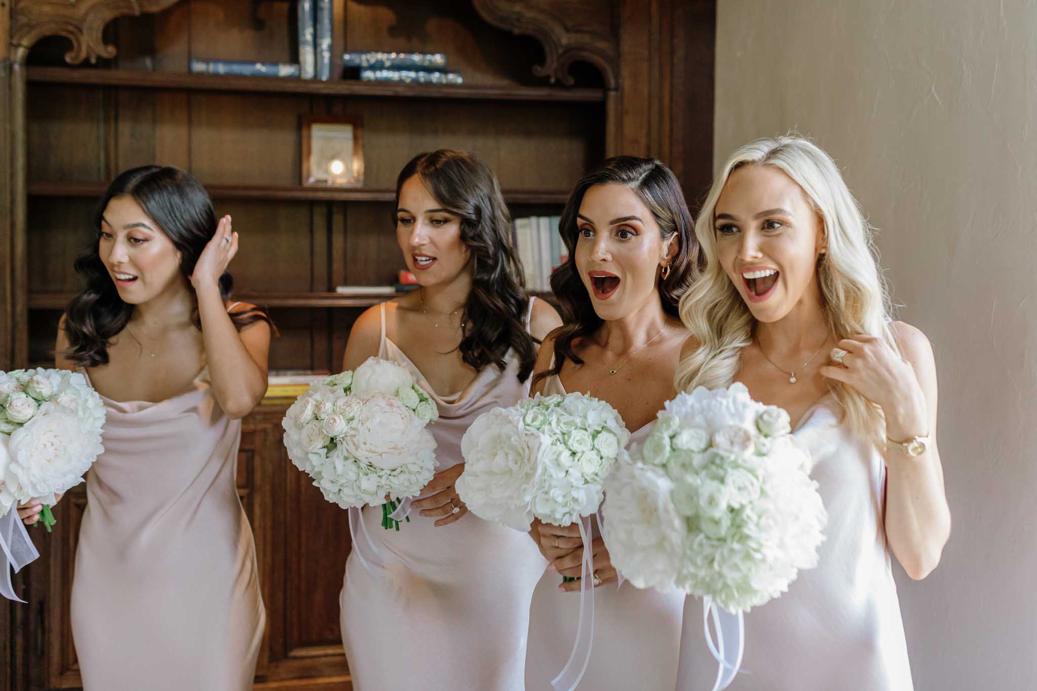 Four bridesmaids in blush cowl-neck dresses laughing together while holding white hydrangea bouquets indoors