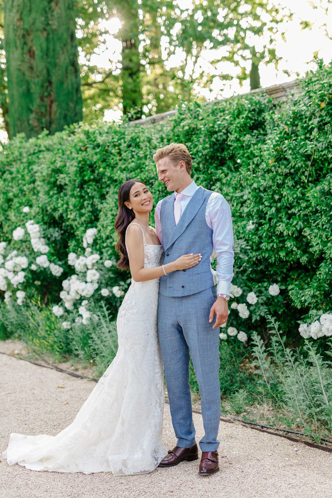 Bride in white lace mermaid gown and groom in slate blue suit on stone path with climbing white roses and ivy