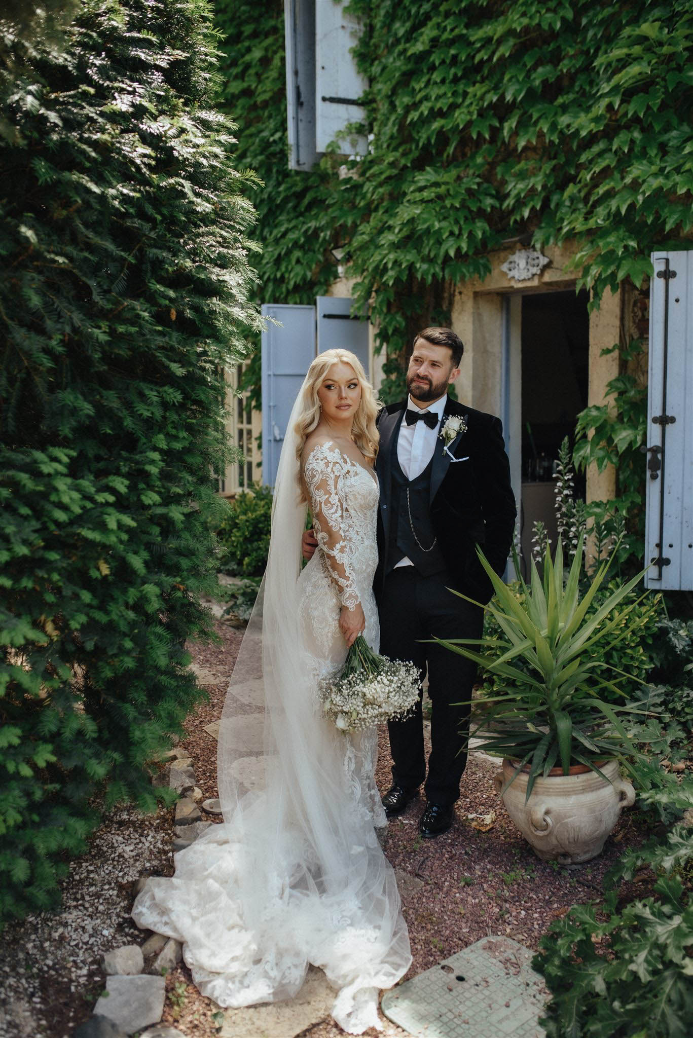 Full-length portrait of bride in lace gown with cathedral veil and groom in black tuxedo on gravel path by ivy-covered sto...
