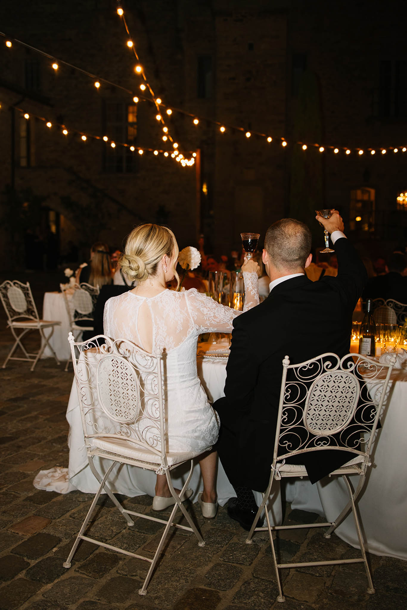 Bride and groom viewed from behind raising glasses in toast at outdoor courtyard reception under string lights at night