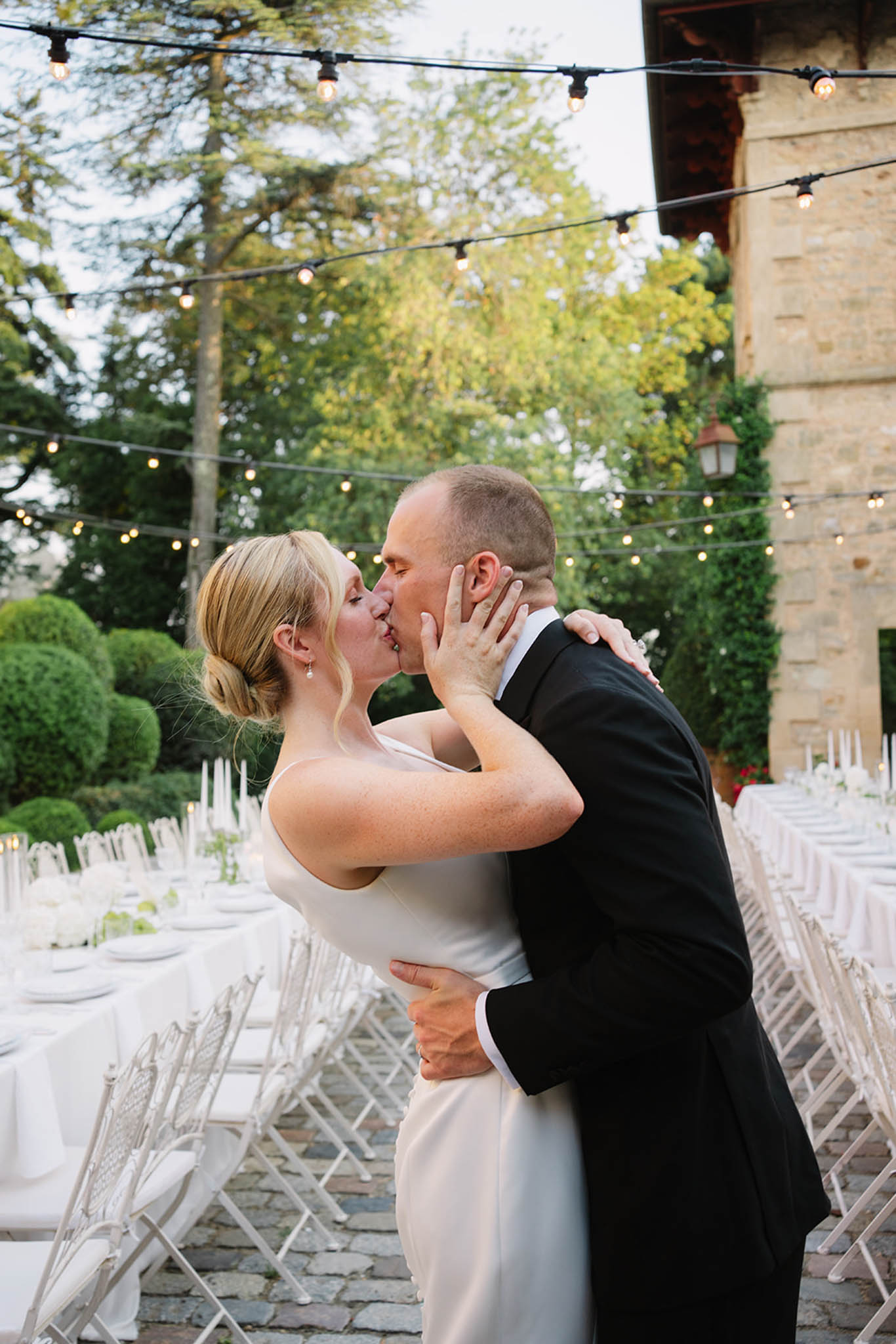 Bride and groom share first kiss in stone courtyard with string lights and white-draped reception tables