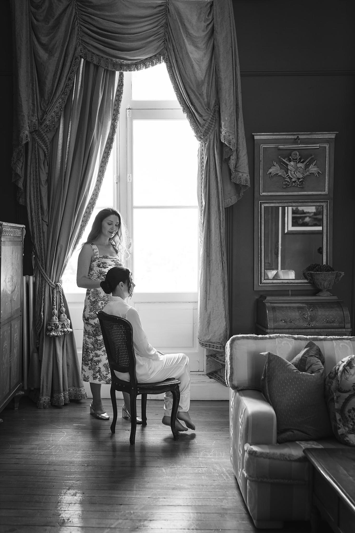 Black and white bridal preparation, bride at tall window, bridesmaid assisting, period furnishings throughout room