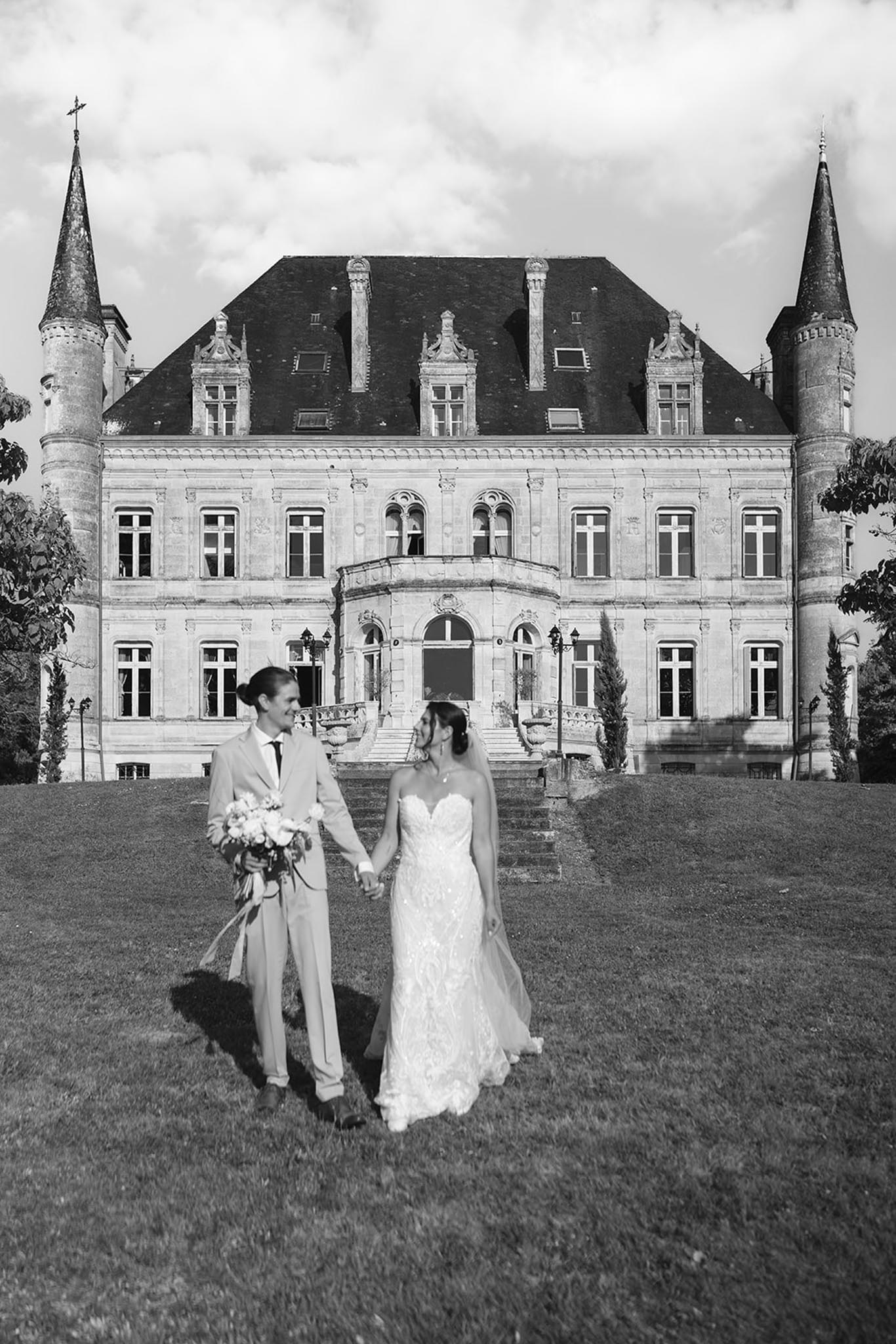 Bride in lace gown and groom hold hands on lawn before grand chateau with conical turrets, black-and-white