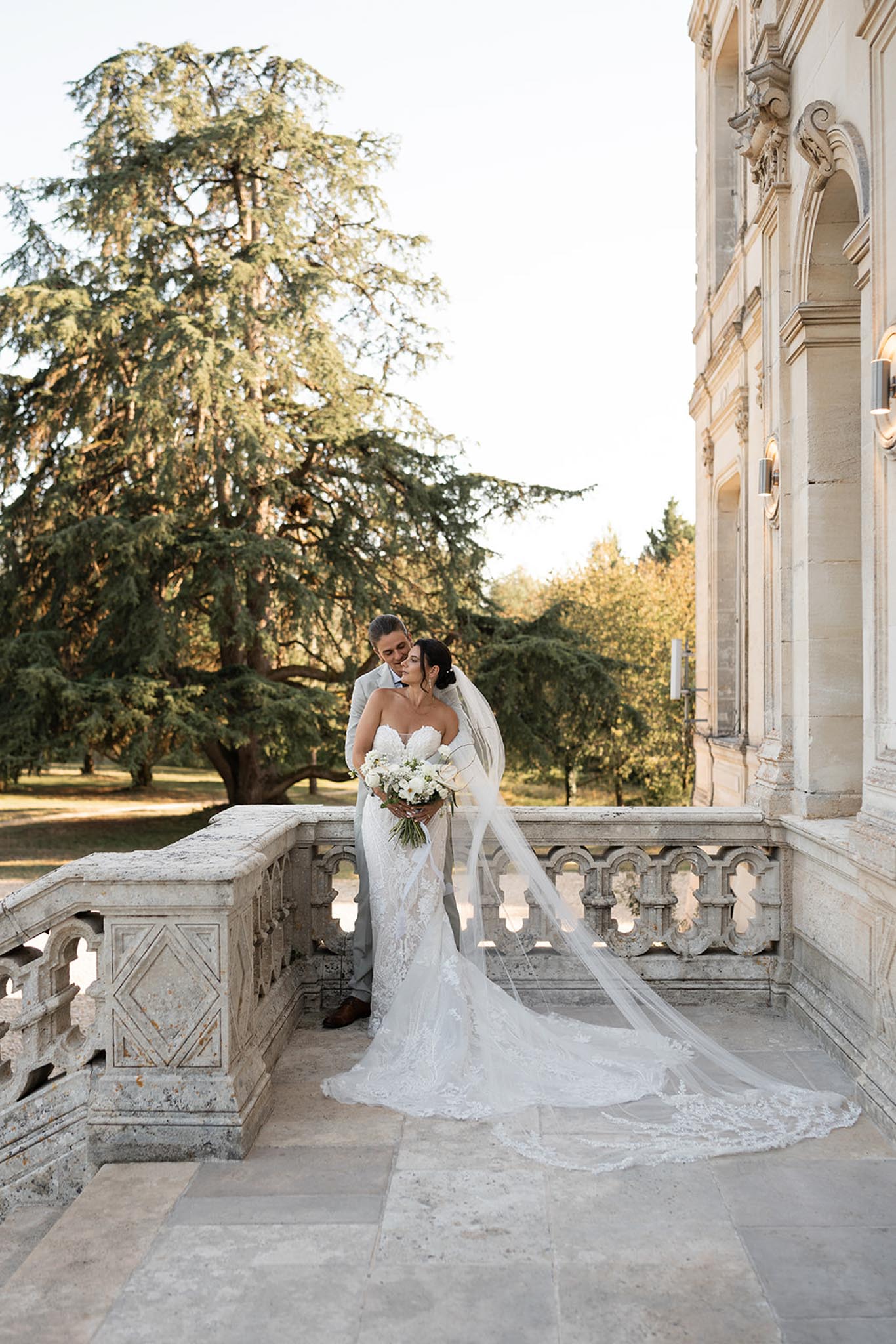 Bride in lace mermaid gown and groom in light blue suit on stone terrace of classical chateau
