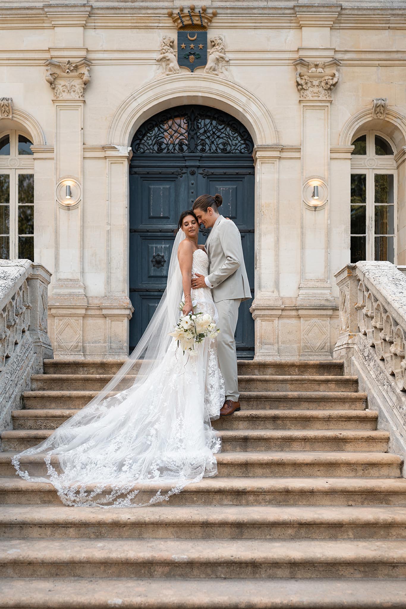 Bride and groom embrace on stone staircase before neoclassical façade with columns and arched blue doorway