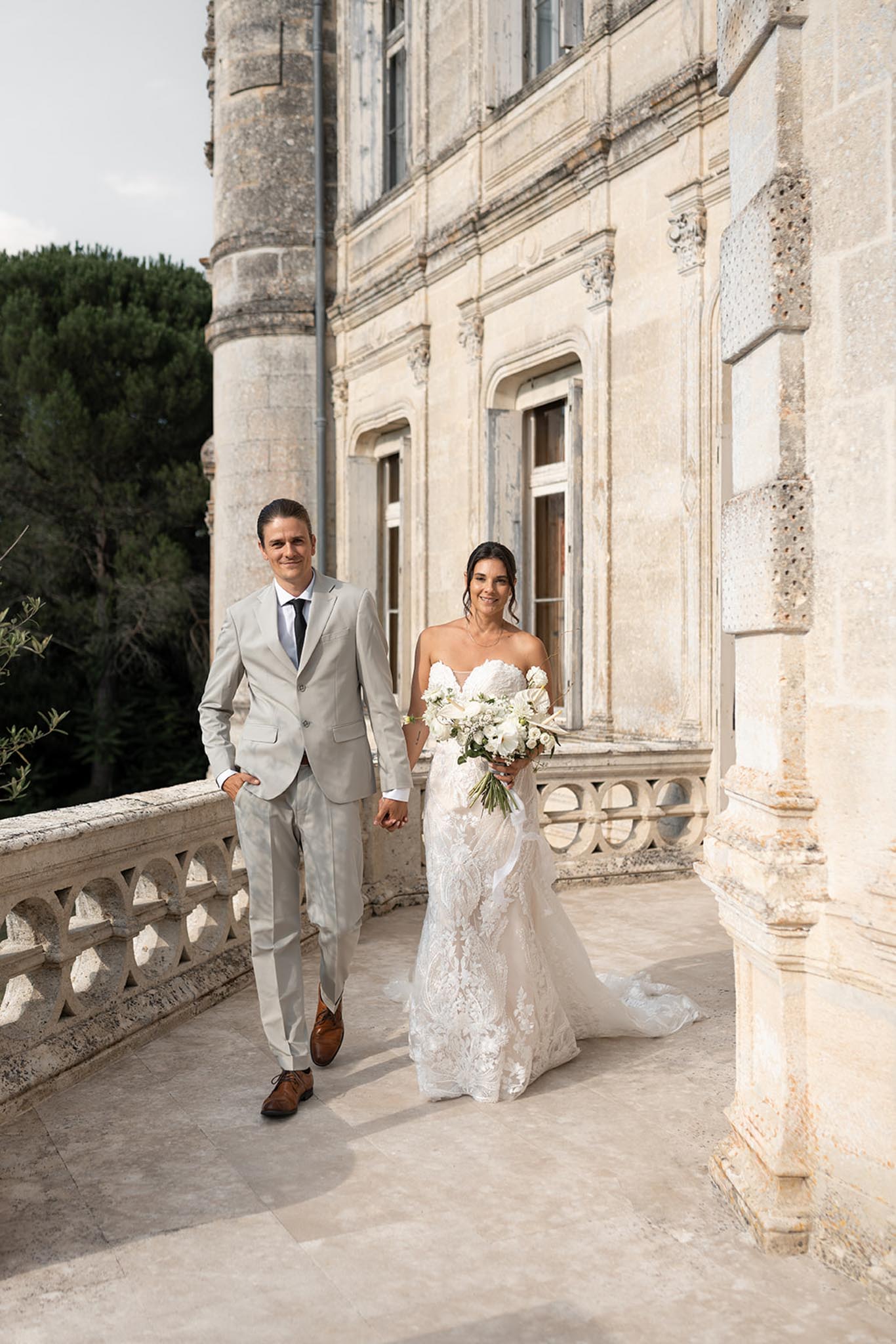 Bride and groom walking hand-in-hand on stone terrace with carved balustrade beside pale limestone château facade