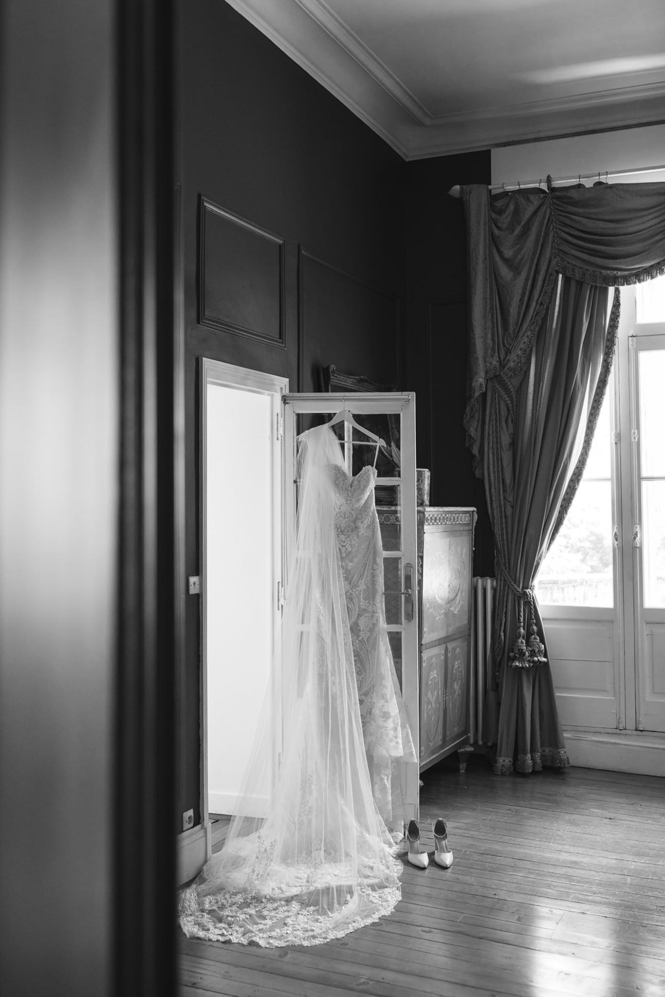 Black and white photo of wedding dress and veil hanging on vintage wardrobe with bridal shoes below