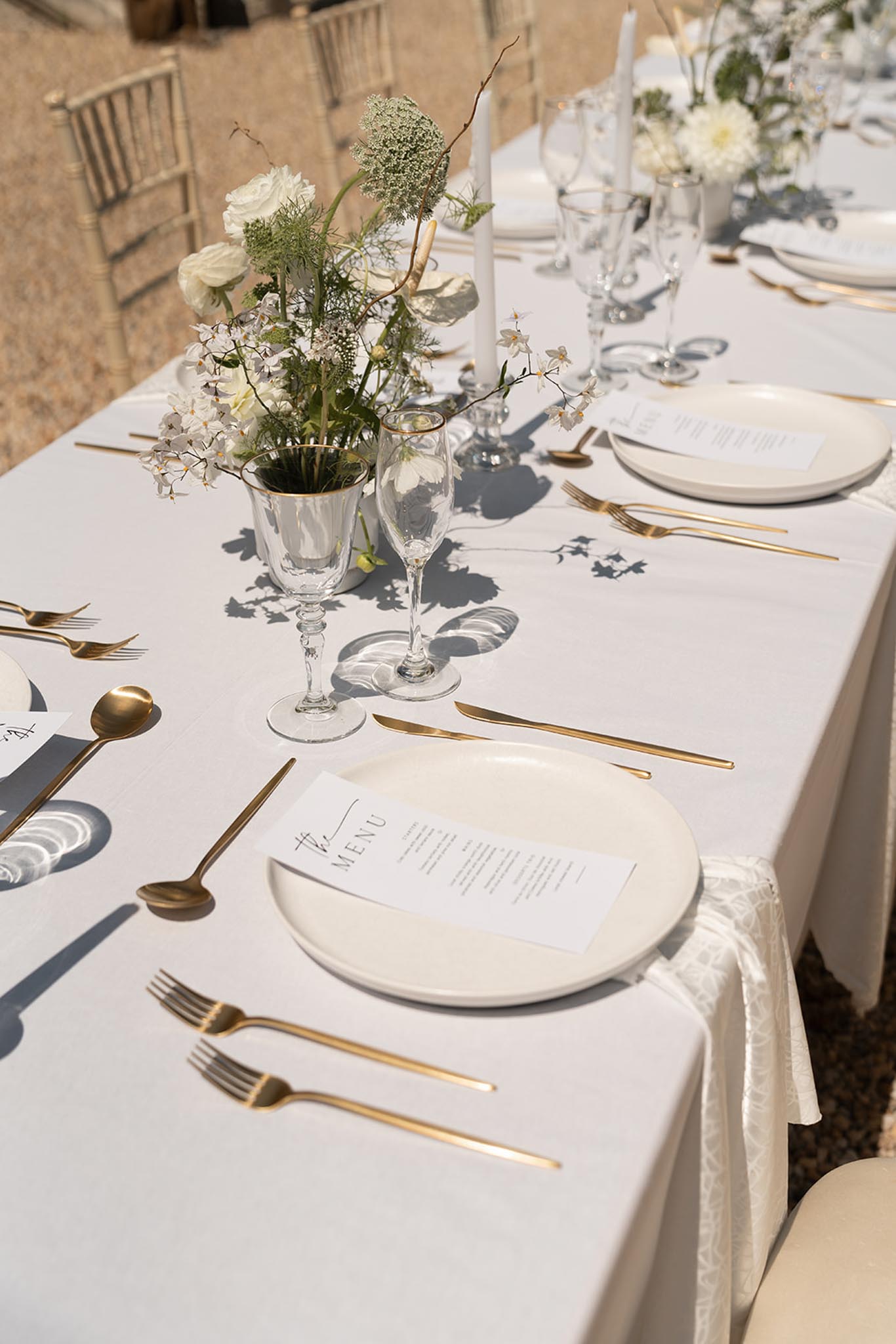 Outdoor reception table with white linens, gold flatware, champagne flutes, and white dahlia centerpieces in bright sunlight