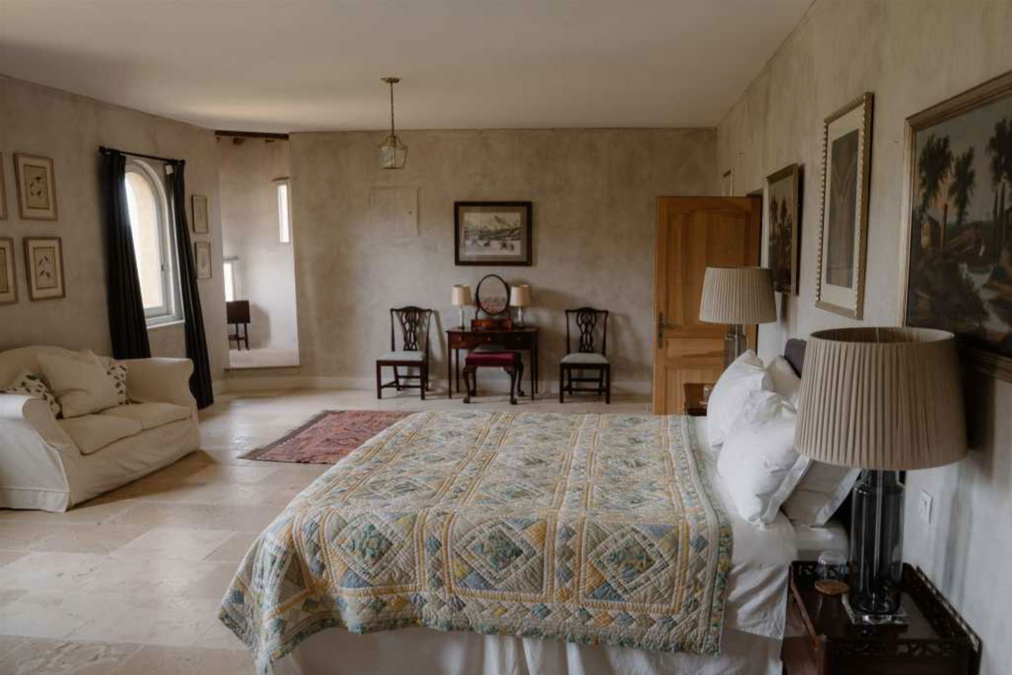 Spacious chateau bedroom with limestone floors, patchwork quilt bed, antique dressing table, and framed artwork