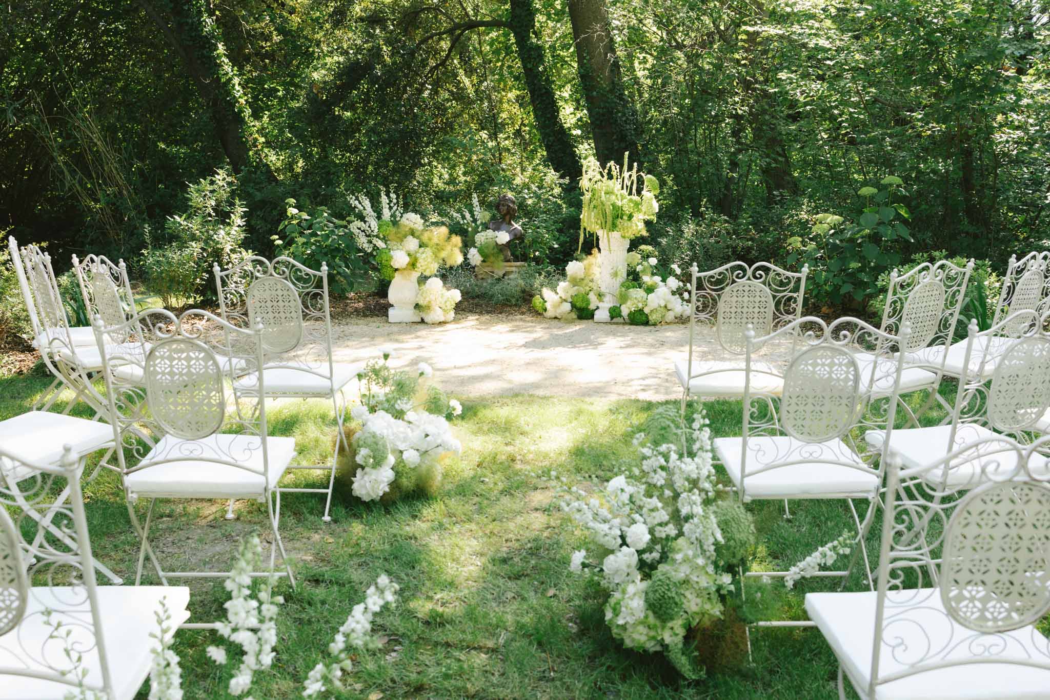 Ceremony setup in woodland garden with white ornate iron chairs and tall white floral altar installation under tree canopy