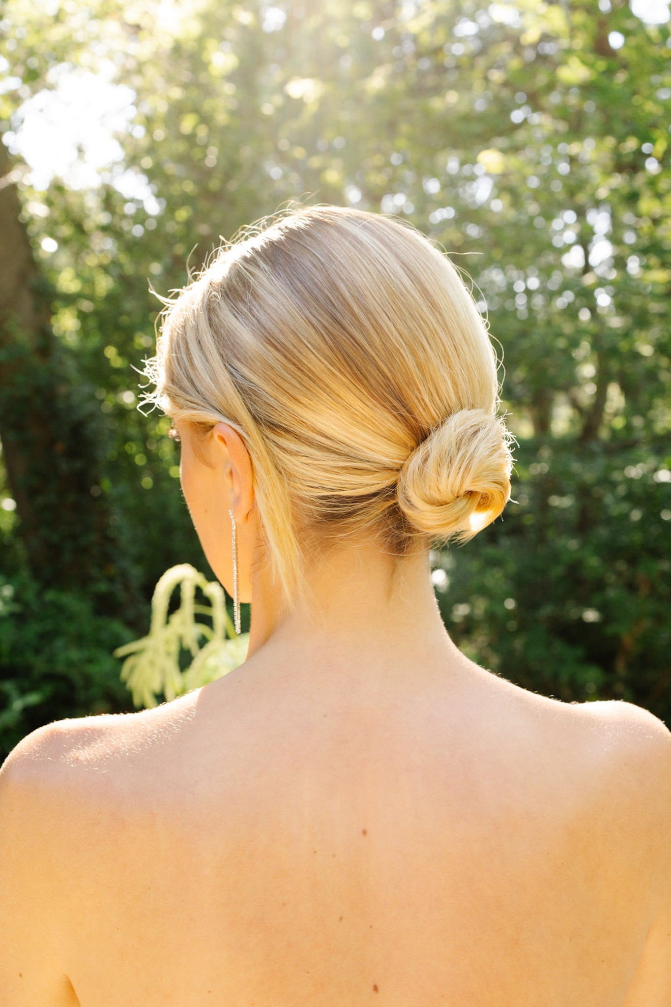 Bride photographed from behind showing low twisted bun hairstyle, chain earrings, and gold dress in a sunlit garden
