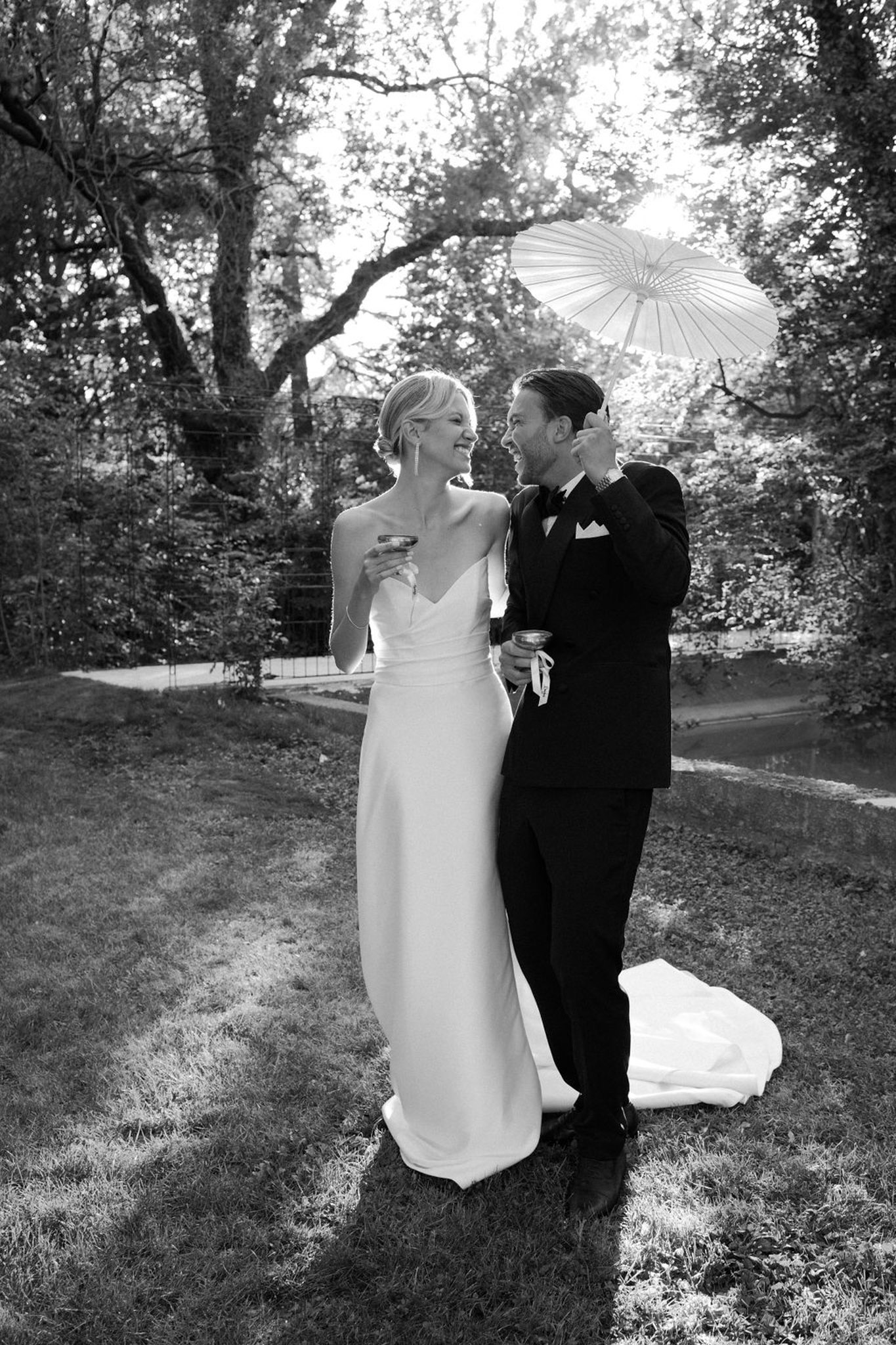 Black-and-white portrait of bride and groom walking arm-in-arm through wooded garden with white parasol